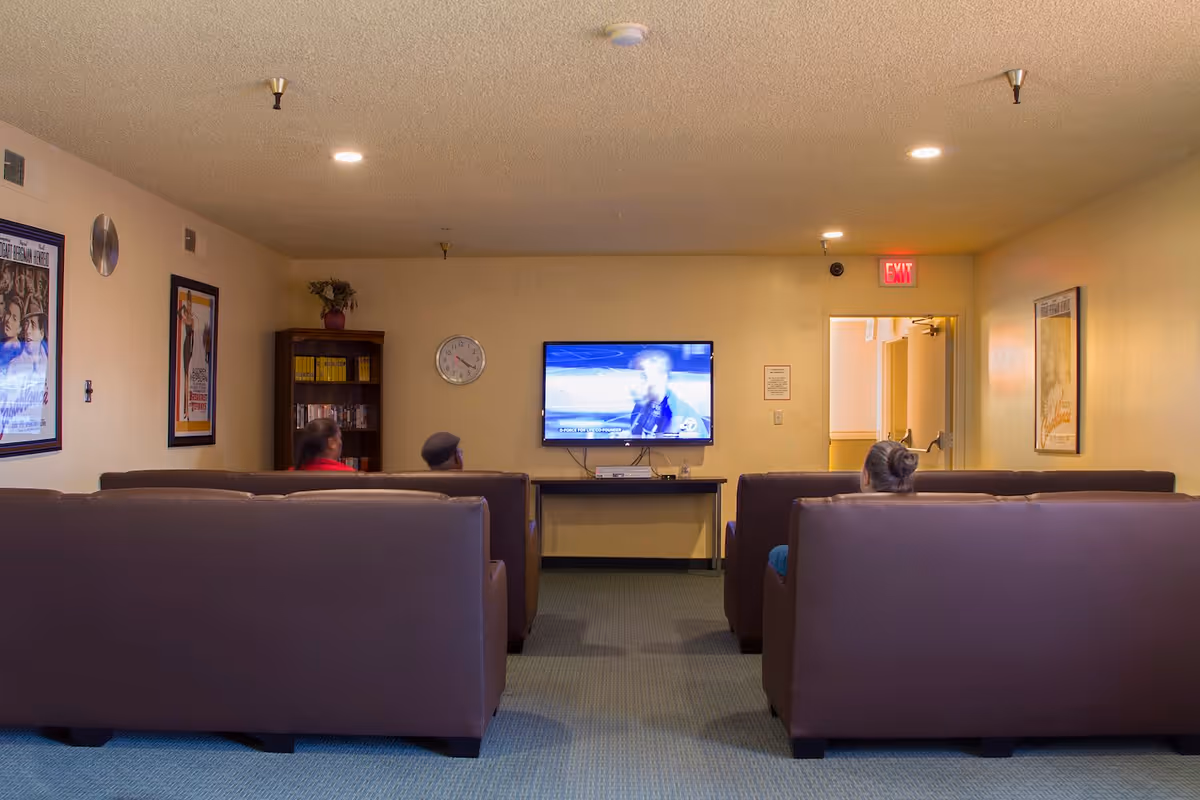 A common area with three brown couches facing a wall-mounted television. Three people are seated on the couches watching TV. The room has beige walls with framed posters, a clock, a bookshelf with books and DVDs, and an exit door with an illuminated exit sign above it.