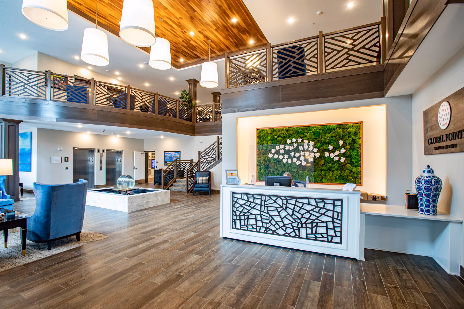 Bright and spacious senior living facility lobby with a modern reception desk featuring a decorative front panel. Behind the desk is a large green wall art with white flowers. To the right, a wooden sign reads 'Global Pointe Senior Living'. The lobby has wooden flooring, blue armchairs, a central water feature, and a staircase leading to an upper level with decorative railings. Large pendant lights hang from a wooden ceiling panel.