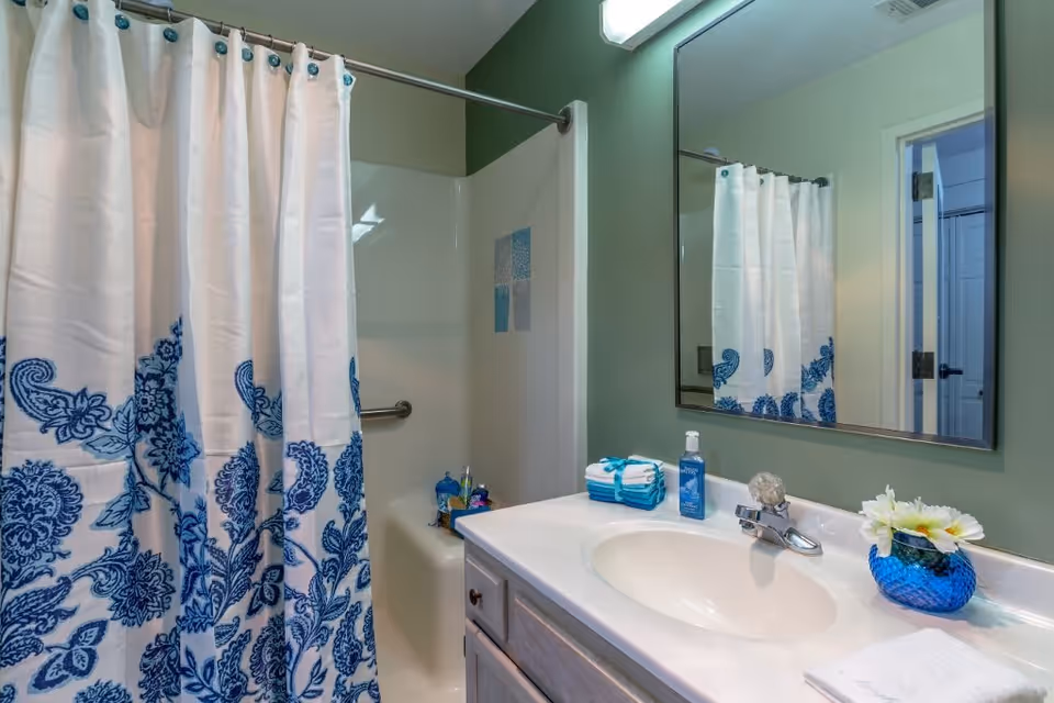 A bathroom with a white shower curtain featuring blue floral patterns, a white sink with a silver faucet, a large mirror above the sink, folded towels, a bottle of hand soap, and a blue vase with white flowers on the countertop.