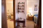 View down a small hallway with a decorative wall shelf and console table, a bathroom visible on the left and a bedroom with a bed and nightstand on the right.