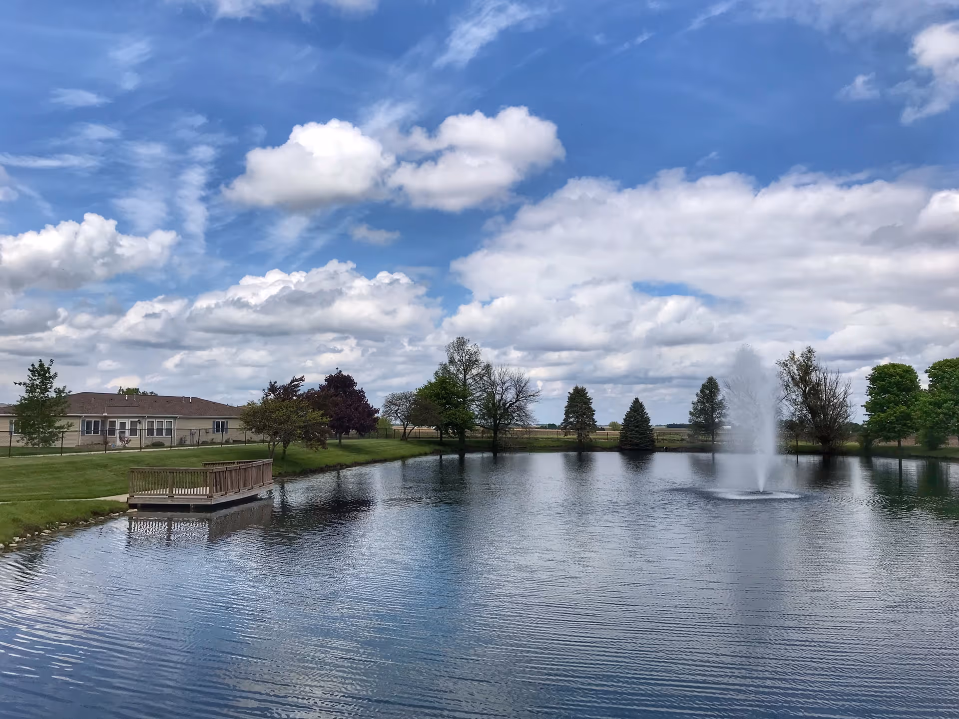 A peaceful outdoor scene at Snyder Village featuring a large pond with a water fountain in the center, surrounded by green grass and various trees under a partly cloudy blue sky. A wooden dock extends into the pond on the left side, and a building is visible in the background.