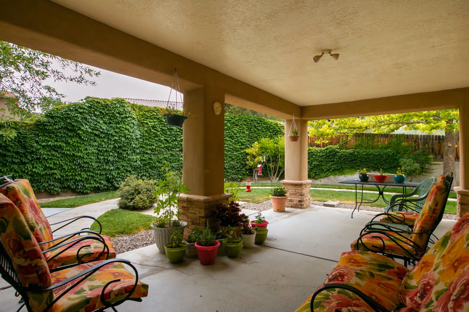Covered outdoor patio area with floral cushioned chairs, potted plants, and a table with chairs. The patio overlooks a green garden with a lawn, bushes, and a wooden fence covered in ivy.
