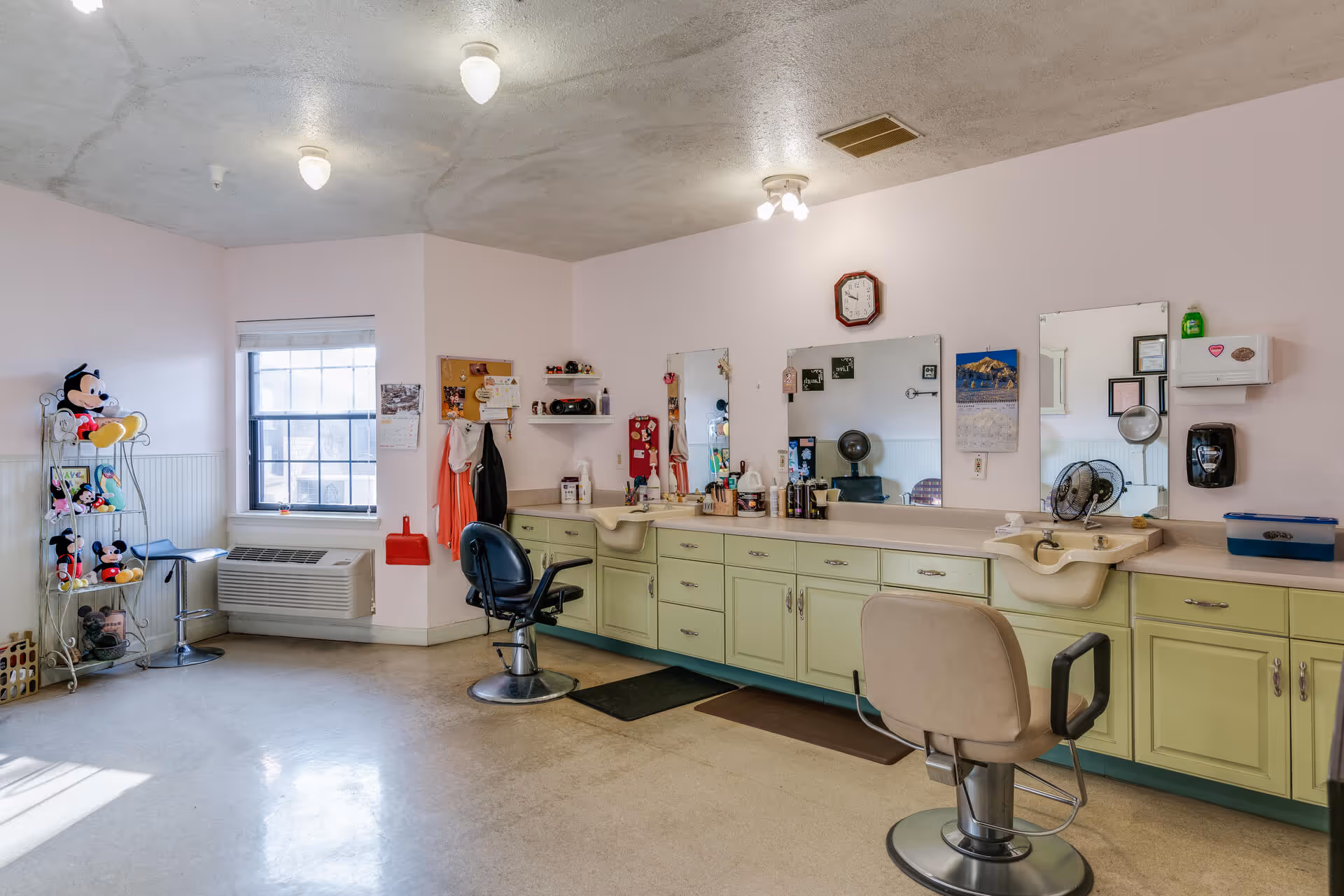 A bright room with pastel green cabinets and two salon chairs in front of a long counter with sinks and mirrors. The room has a window with blinds, a wall-mounted air conditioning unit, and a rack with various stuffed Mickey Mouse toys. The walls are light pink, and there are ceiling lights illuminating the space.