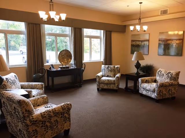 A cozy living room with four floral-patterned armchairs arranged around a small dark wood side table with a lamp. There are two large windows with brown curtains letting in natural light. A dark wood console table with decorative items is placed against the wall beneath the windows. Two abstract paintings hang on the wall, and two ceiling light fixtures illuminate the room.