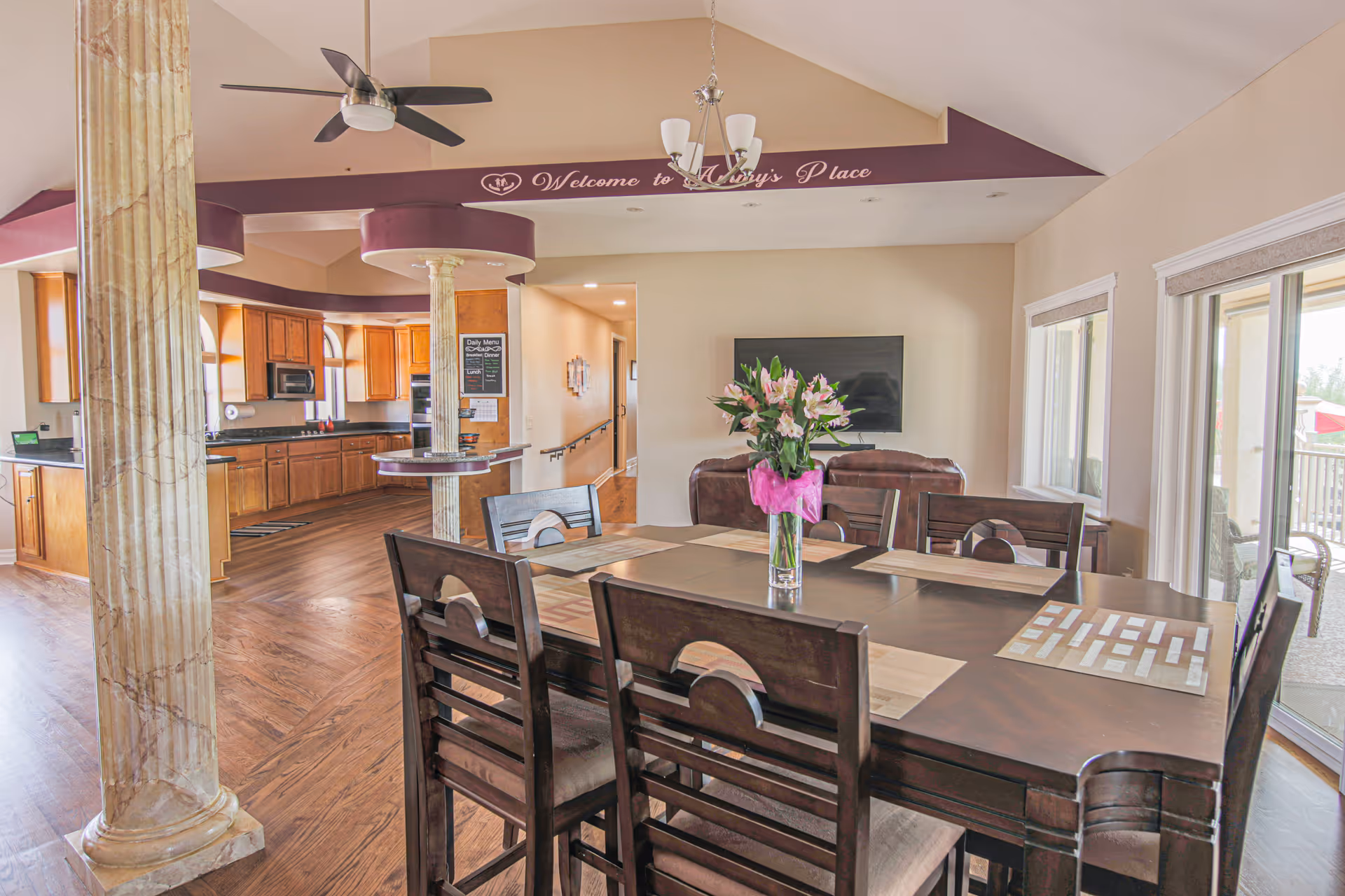 Open dining area with a large wooden table and vase of flowers, adjacent kitchen, living room seating, and sliding glass doors.