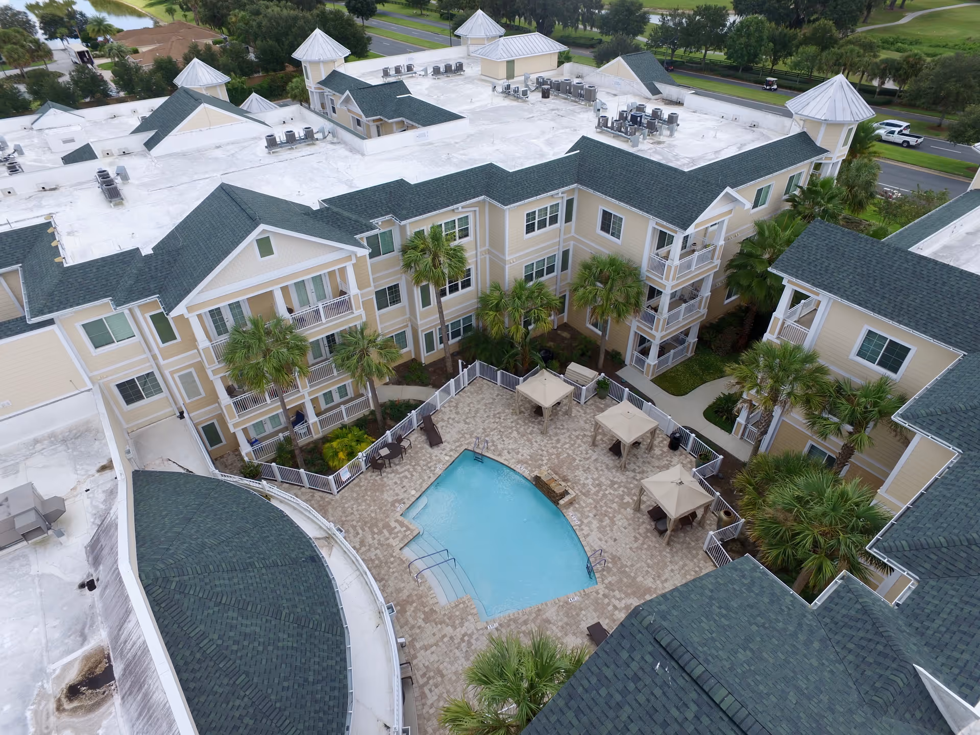 Aerial view of a senior living complex courtyard with a central swimming pool, surrounding three-story beige buildings, palm trees, and poolside cabanas.