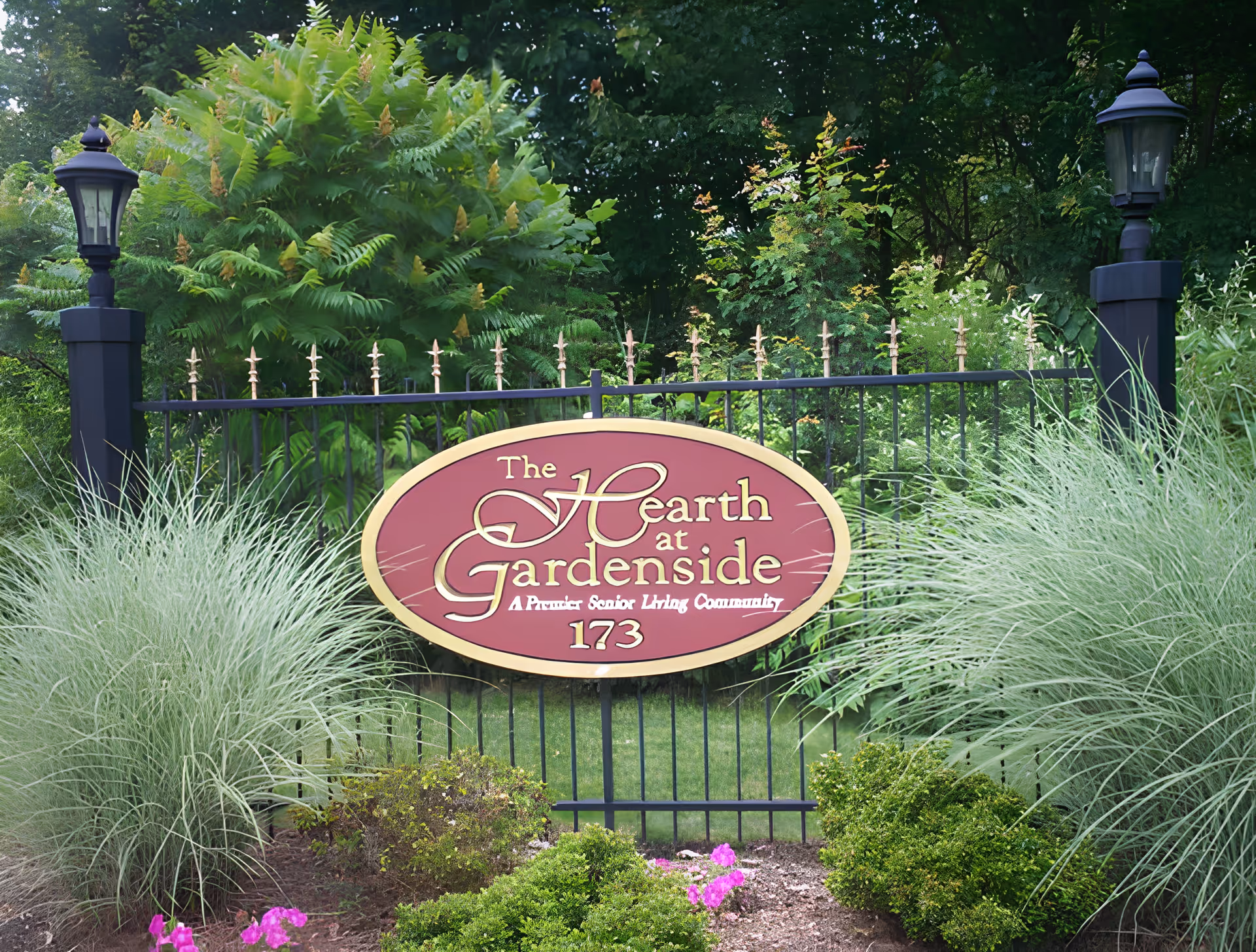 Entrance sign for "The Hearth at Gardenside" mounted on a black wrought-iron fence surrounded by ornamental grasses, shrubs, and lantern posts.