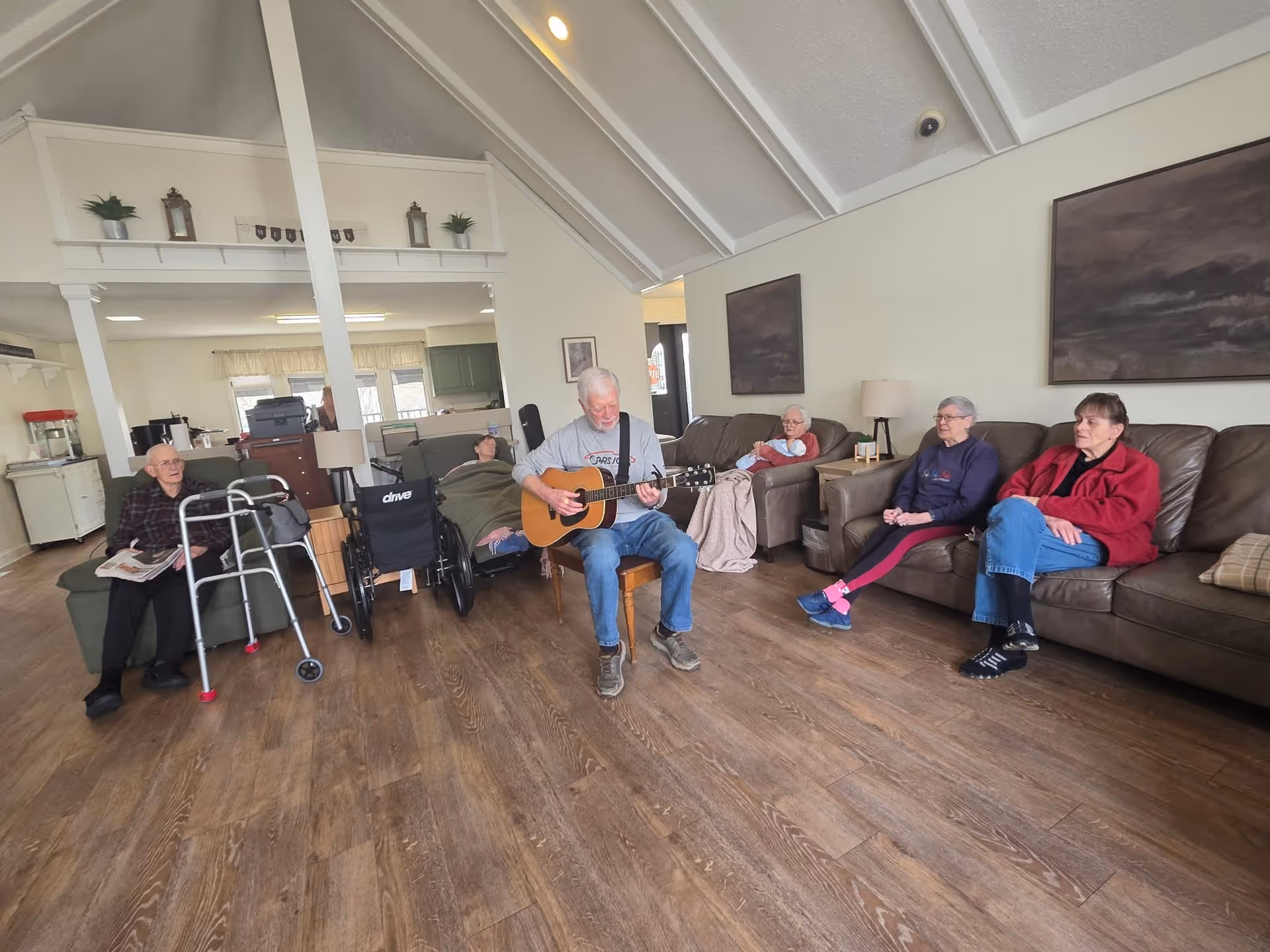 Several elderly residents seated in a common living room area while a man plays an acoustic guitar.