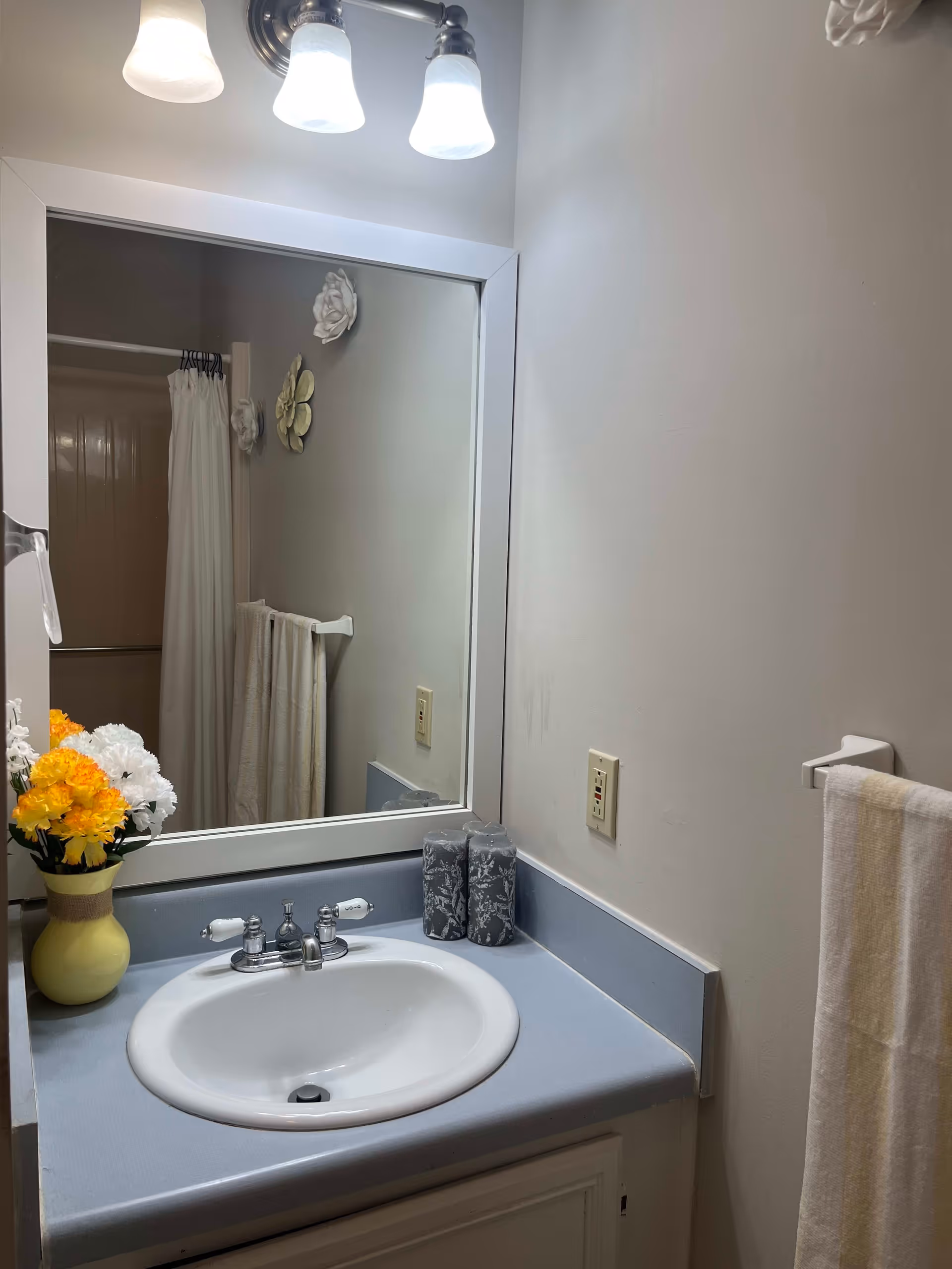 A bathroom sink with a light blue countertop, a white oval basin, and a silver faucet. Above the sink is a large rectangular mirror with a white frame, reflecting a shower curtain and decorative wall flowers. To the left of the sink is a yellow vase with yellow and white flowers. Two gray patterned containers are placed on the countertop to the right of the sink. A beige towel hangs on a towel rack mounted on the wall to the right. The wall is painted light gray, and there is a light fixture with three bulbs above the mirror.