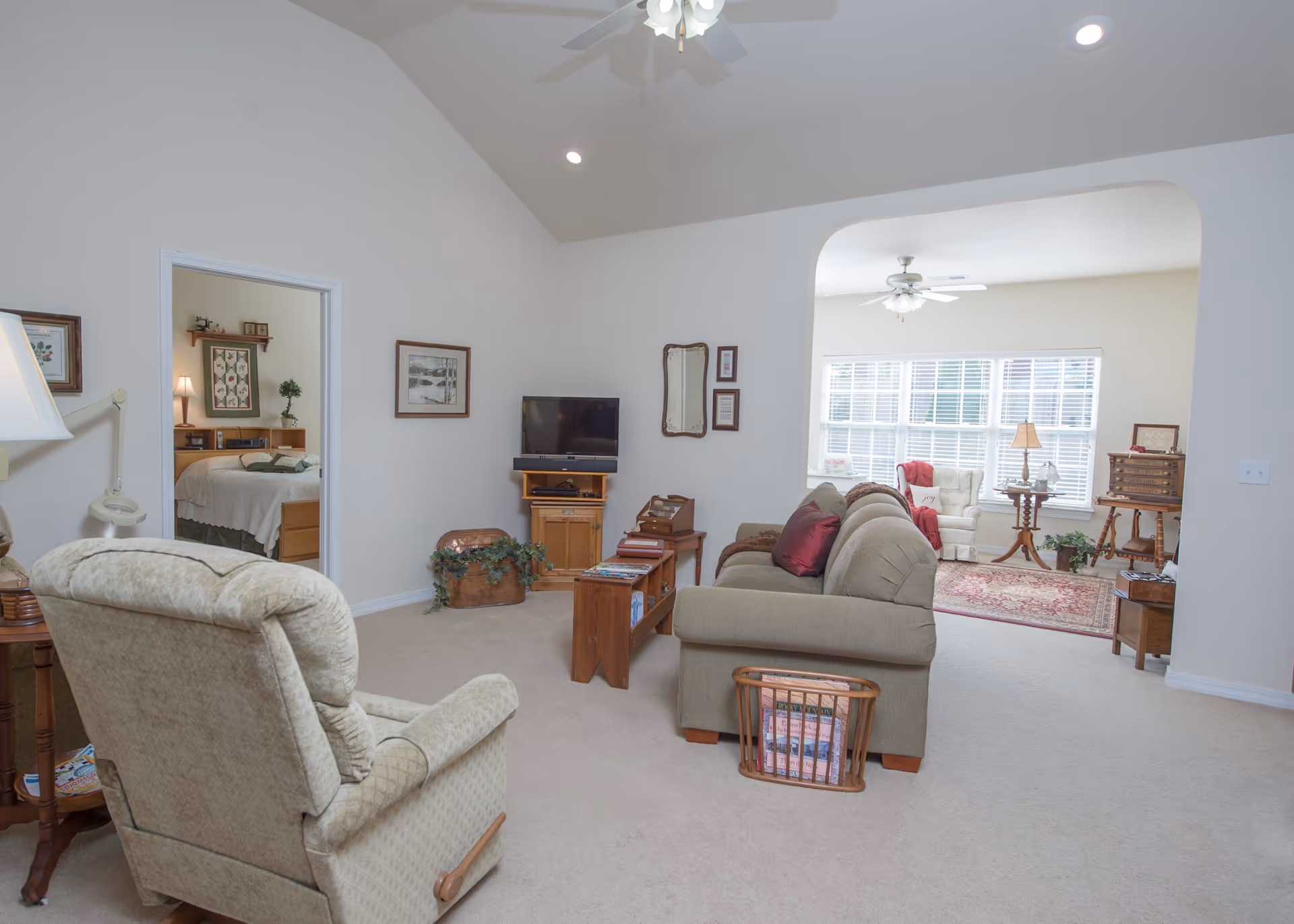 Bright living room with a sofa, recliner, TV and wooden side tables, opening via an archway to a sunlit sitting area.