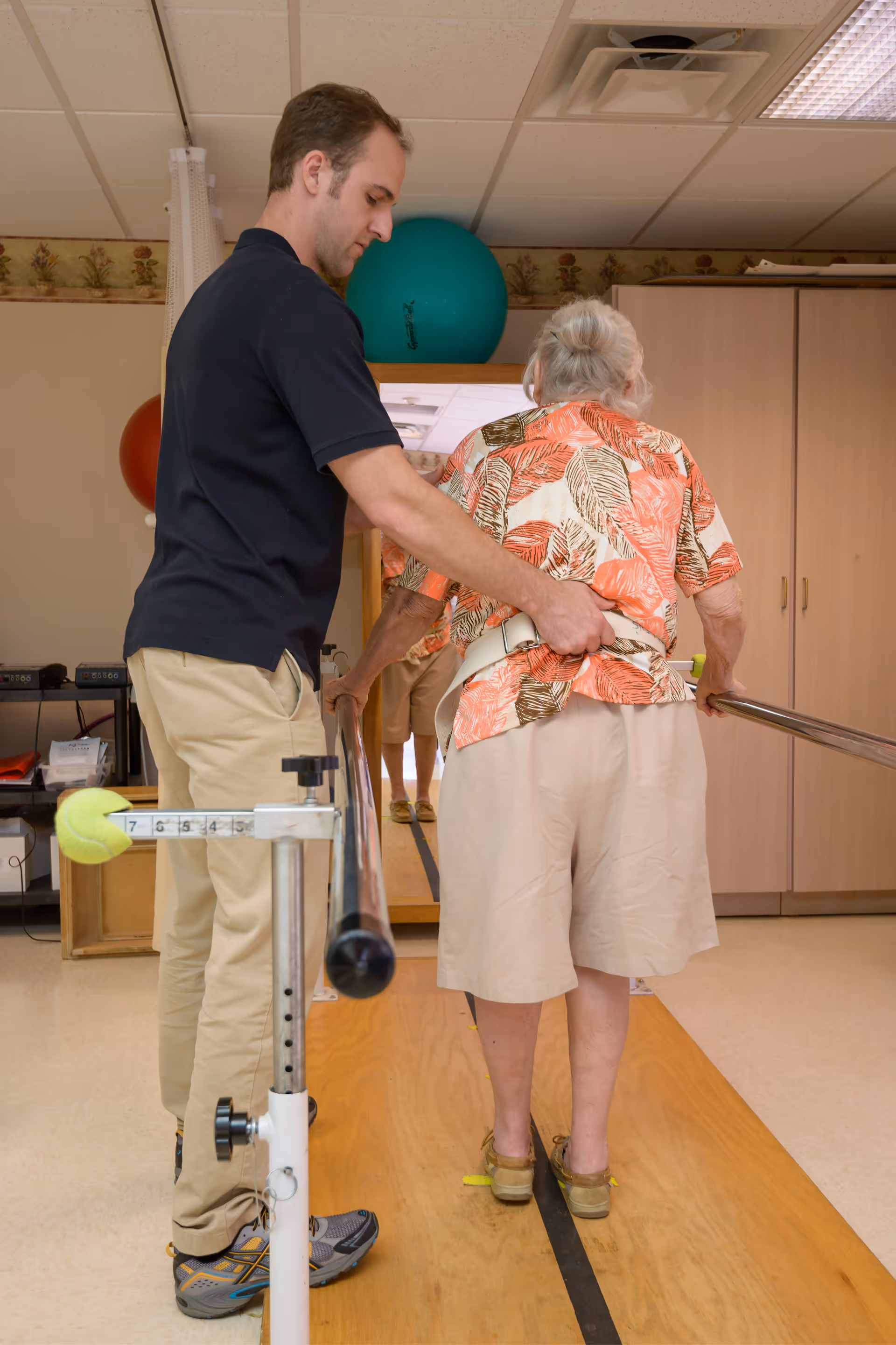 A male caregiver assists an elderly woman with white hair as she walks between parallel bars in a rehabilitation or physical therapy room. The woman is wearing a floral shirt and beige shorts, and the caregiver is supporting her by holding her waist. The room has wooden flooring for walking exercises and exercise balls in the background.