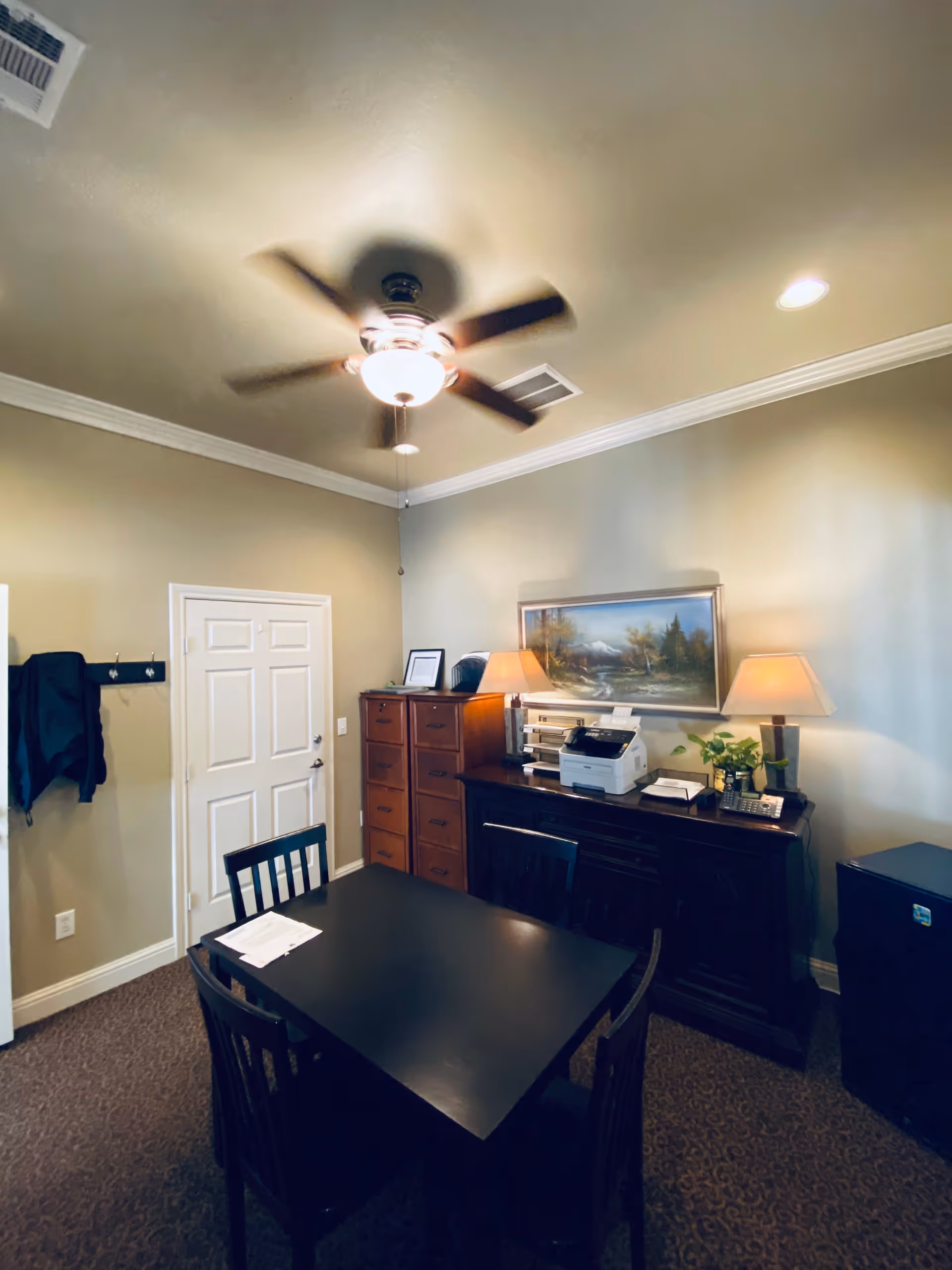 Interior room with a ceiling fan, a four-seat black table, filing cabinets, a printer, lamps, and a framed landscape painting.