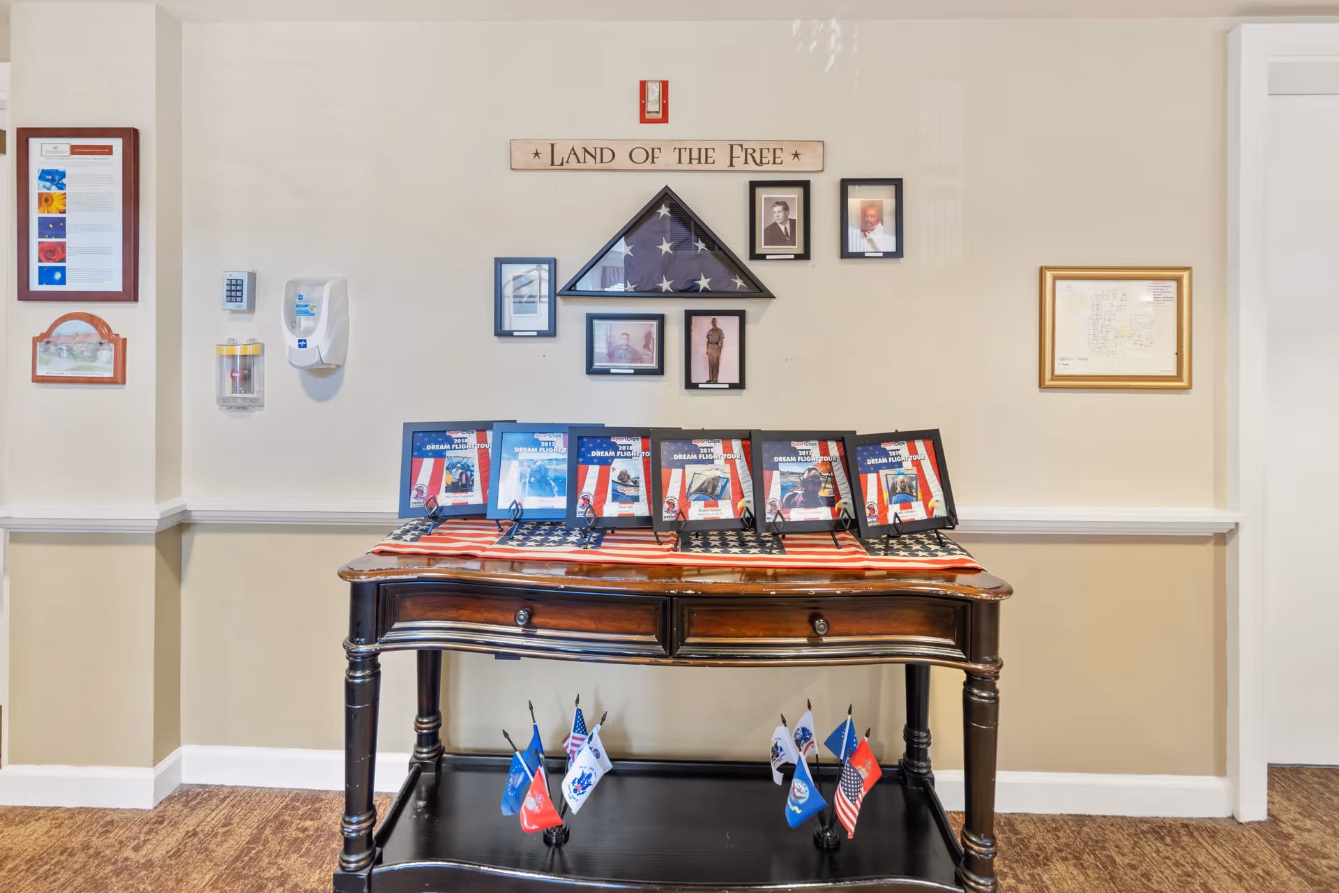 A patriotic display in a senior living facility featuring a wooden table draped with an American flag, several framed certificates or awards, and small flags on the lower shelf. Above the table is a folded American flag in a triangular case, surrounded by framed photographs and a wooden sign that reads 'LAND OF THE FREE'. The walls are beige with white trim, and there are hand sanitizer and other wall-mounted dispensers on the left side.