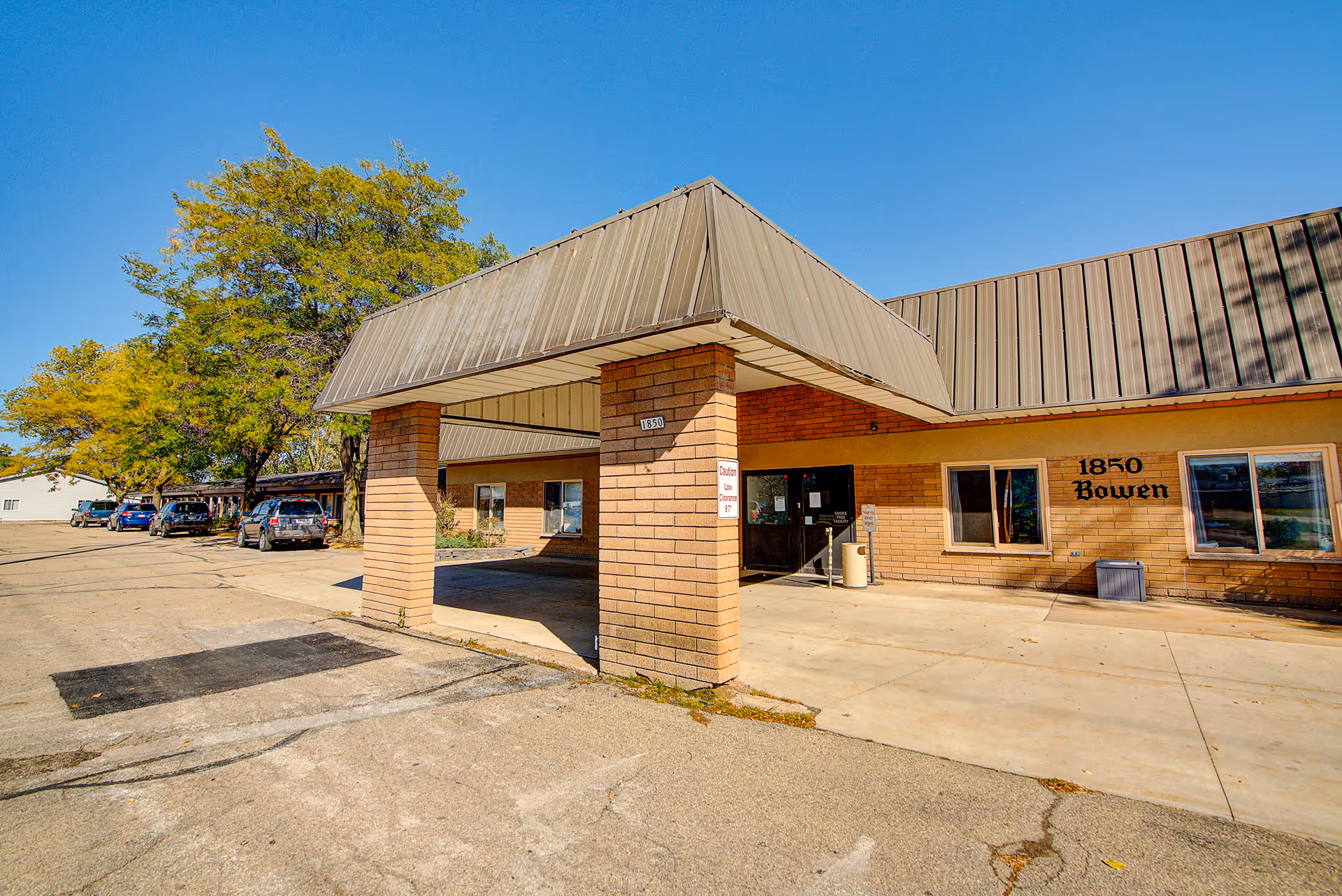 Exterior view of a single-story brick building with a covered entrance supported by two brick pillars. The building has a metal roof and the address number 1850 is visible on the pillar and on the building wall next to the entrance. Several cars are parked along the driveway, and trees with green and yellow leaves are visible under a clear blue sky.