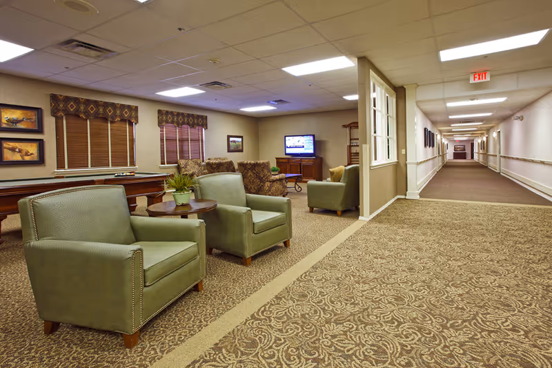 A cozy common area in an assisted living facility with green armchairs, a round wooden table with a potted plant, patterned carpet, a pool table, and a TV mounted on a wooden cabinet. The area opens into a long, well-lit hallway with handrails and framed pictures on the walls.