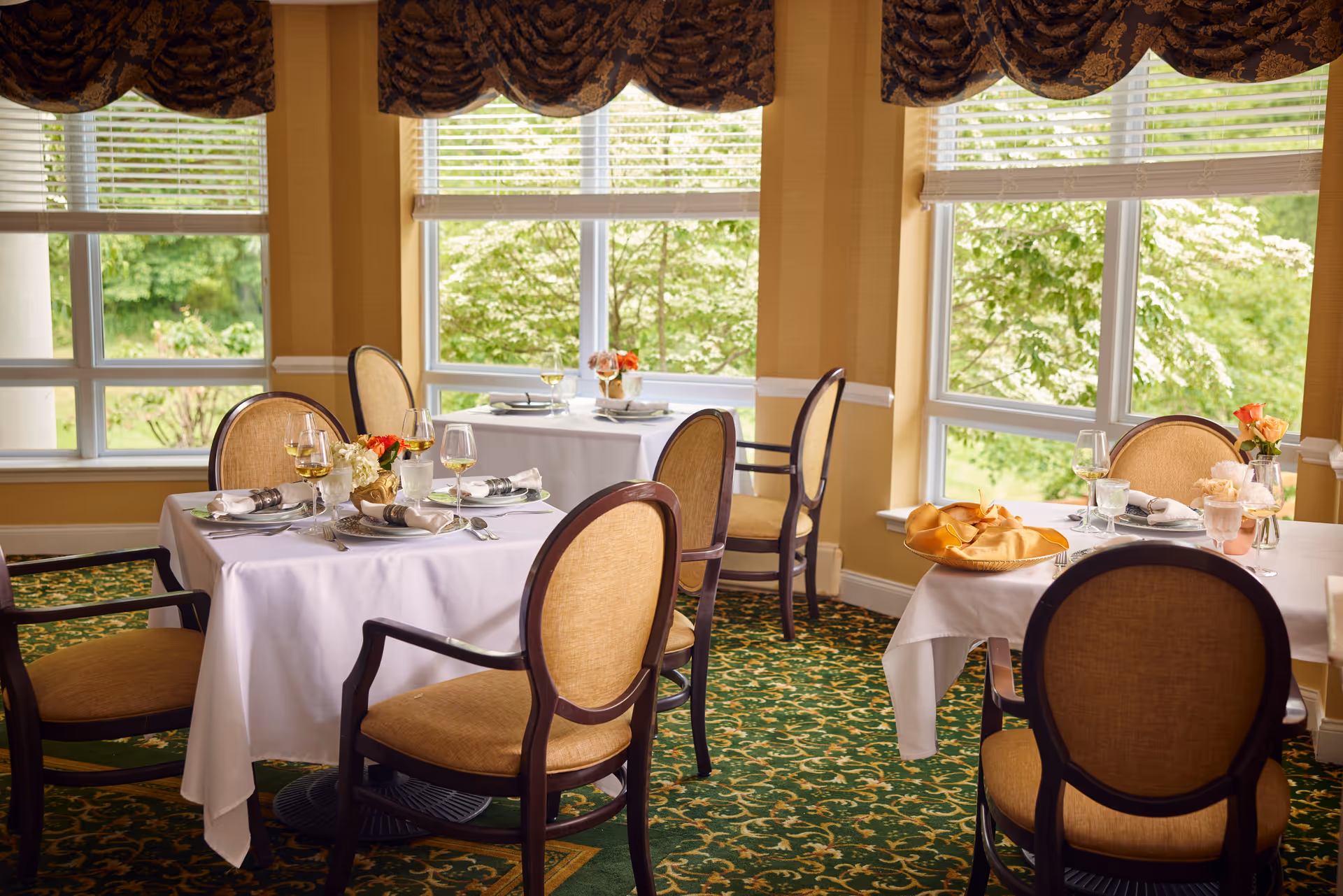 A bright dining room with large windows showing green trees outside. The room has tables covered with white tablecloths, set with plates, glasses, napkins, and small flower arrangements. The chairs have wooden frames with beige upholstered backs and seats. The carpet has a green and gold pattern, and the windows have dark patterned valances.