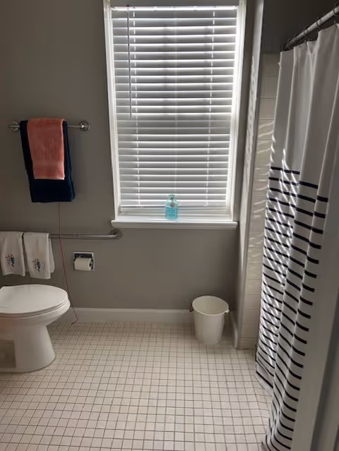 Bathroom with a toilet, window with closed blinds, a striped shower curtain, towels on racks, and a tiled floor.