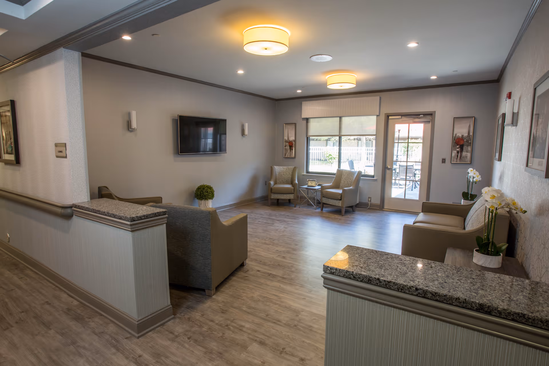 A spacious and well-lit common area in a senior living facility with comfortable armchairs, a wall-mounted flat screen TV, large windows, and a glass door leading outside. The room features wood flooring, neutral-colored walls, modern ceiling lights, and decorative plants and artwork.