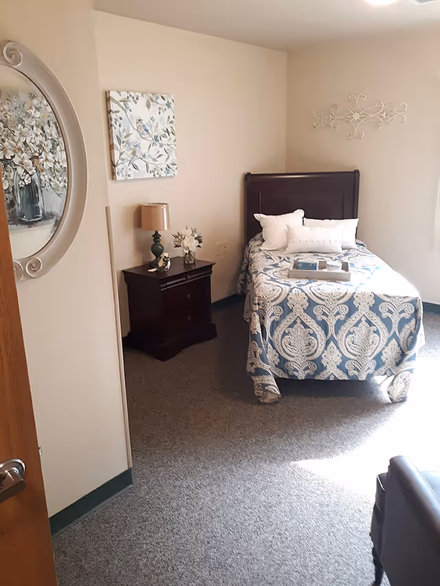 A small, cozy bedroom with a single bed covered in a blue and white patterned bedspread. The bed has several white pillows and a tray with items on it. Next to the bed is a dark wooden nightstand with a green lamp and a small vase of flowers. The walls are light-colored and decorated with a round floral painting and a square bird-themed artwork. The room has carpeted flooring and is softly lit by natural light coming from a window on the right.