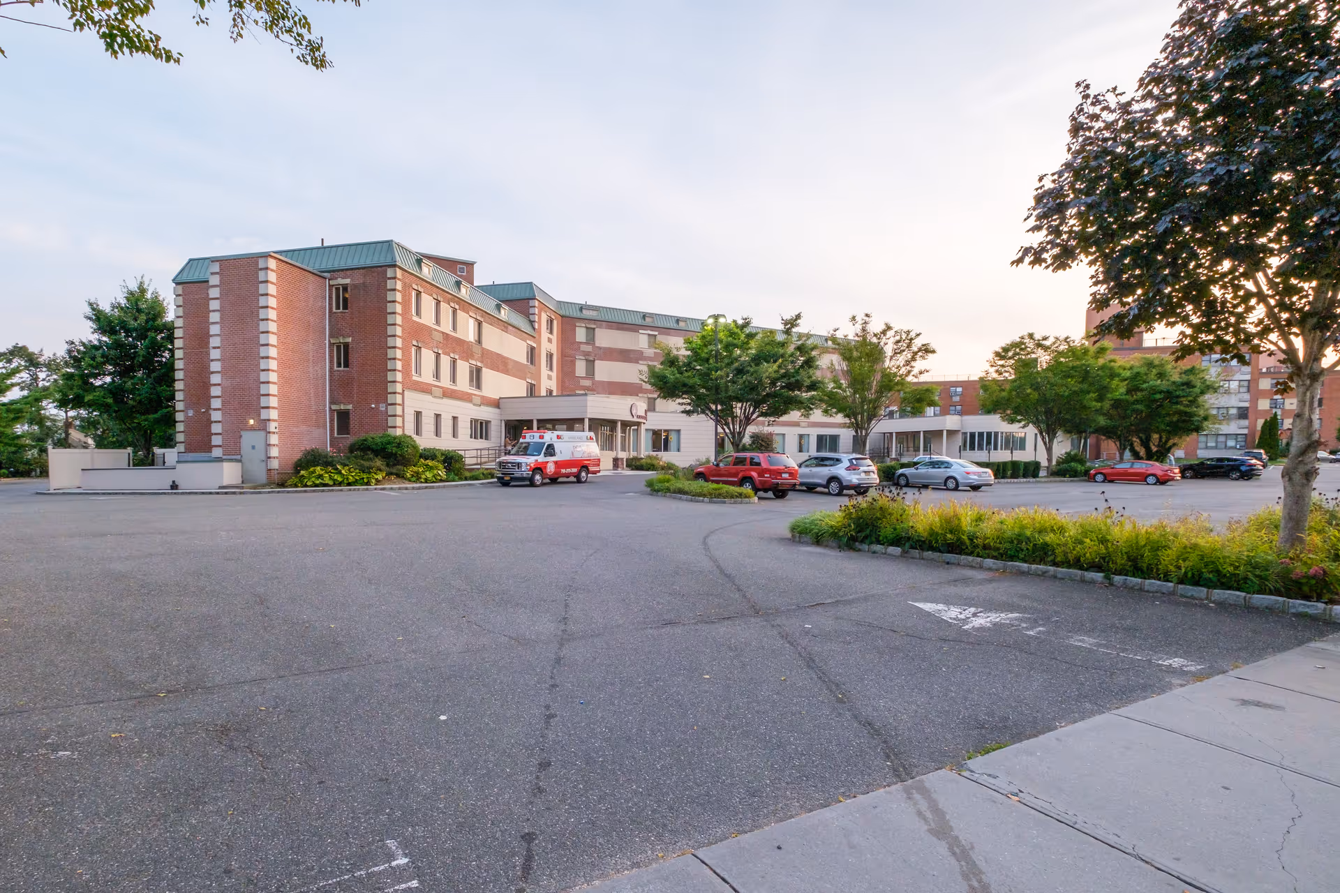 Exterior view of a multi-story brick assisted living facility with a parking lot in front. Several cars and an ambulance are parked near the entrance. Trees and landscaped greenery surround the building under a partly cloudy sky at dusk.