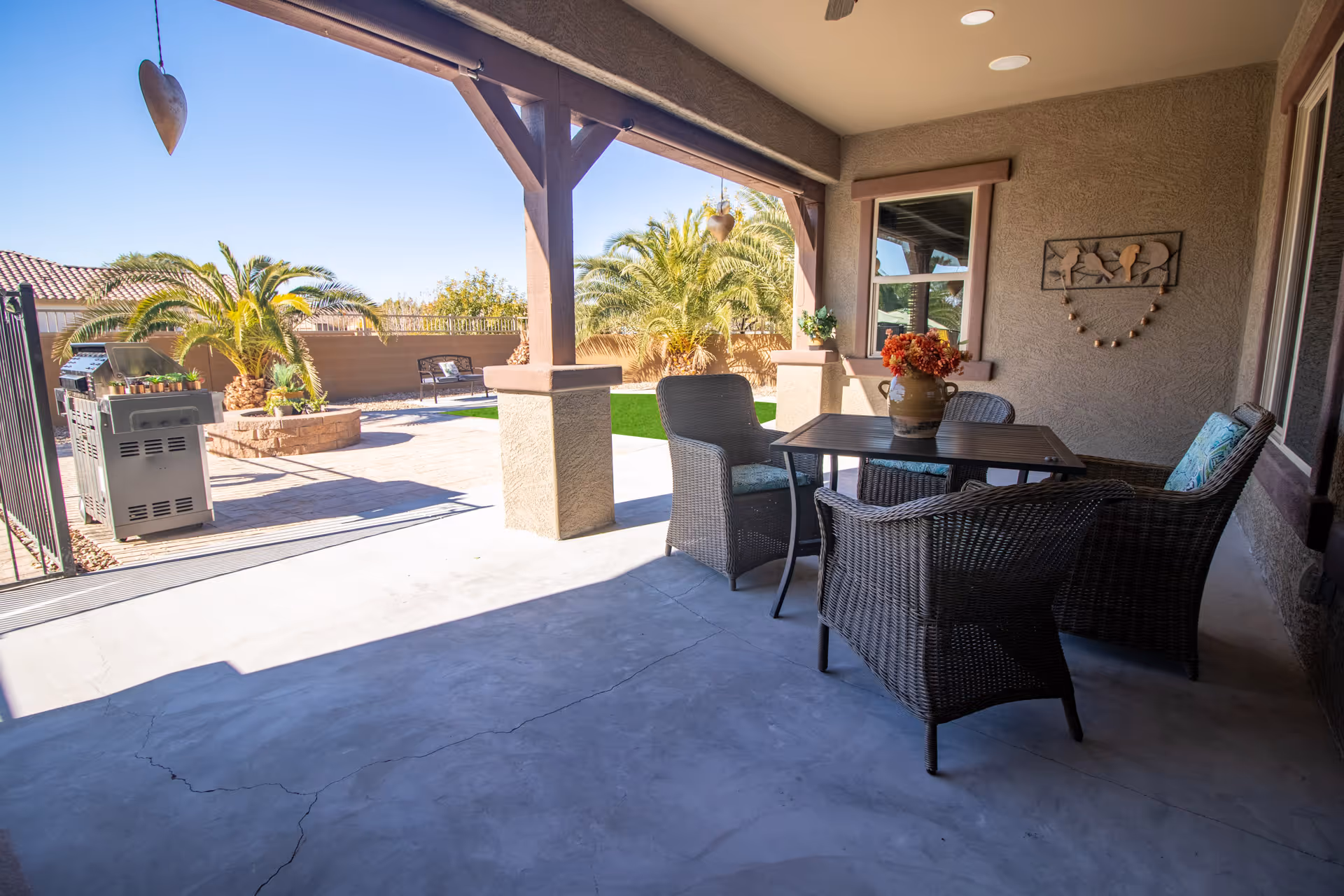 Covered patio with a wicker dining set and vase of flowers overlooking a landscaped backyard with palm trees and a grill.