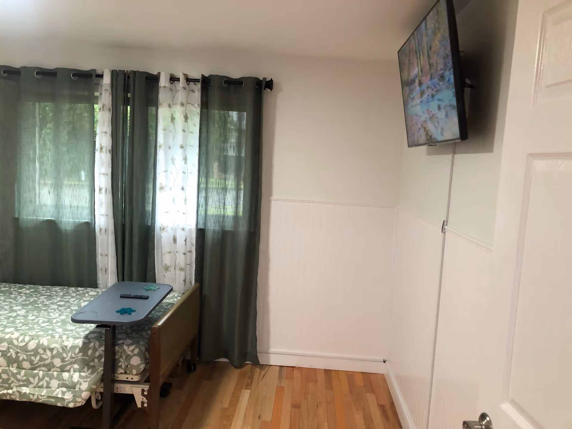 A bedroom with a hospital-style bed covered in green and white leaf-patterned bedding. There is a small overbed table with a remote control and some small objects on it. The room has wooden flooring, white walls with wainscoting, and a window with green and white floral curtains. A flat-screen TV is mounted on the wall opposite the bed.