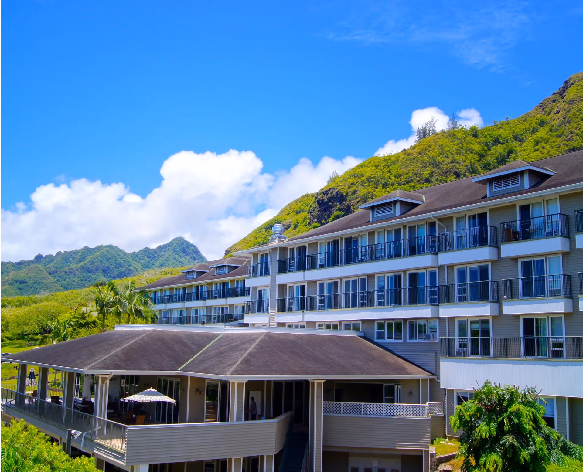 Multi-story senior living building with balconies and a covered porch set against green hills and a bright blue sky.
