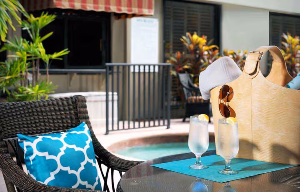 Outdoor seating area with a wicker chair featuring a blue and white patterned cushion, a glass table with two glasses of water garnished with lemon slices, and a beige tote bag holding a hat and sunglasses. In the background, there are plants, a railing, and part of a building with windows.