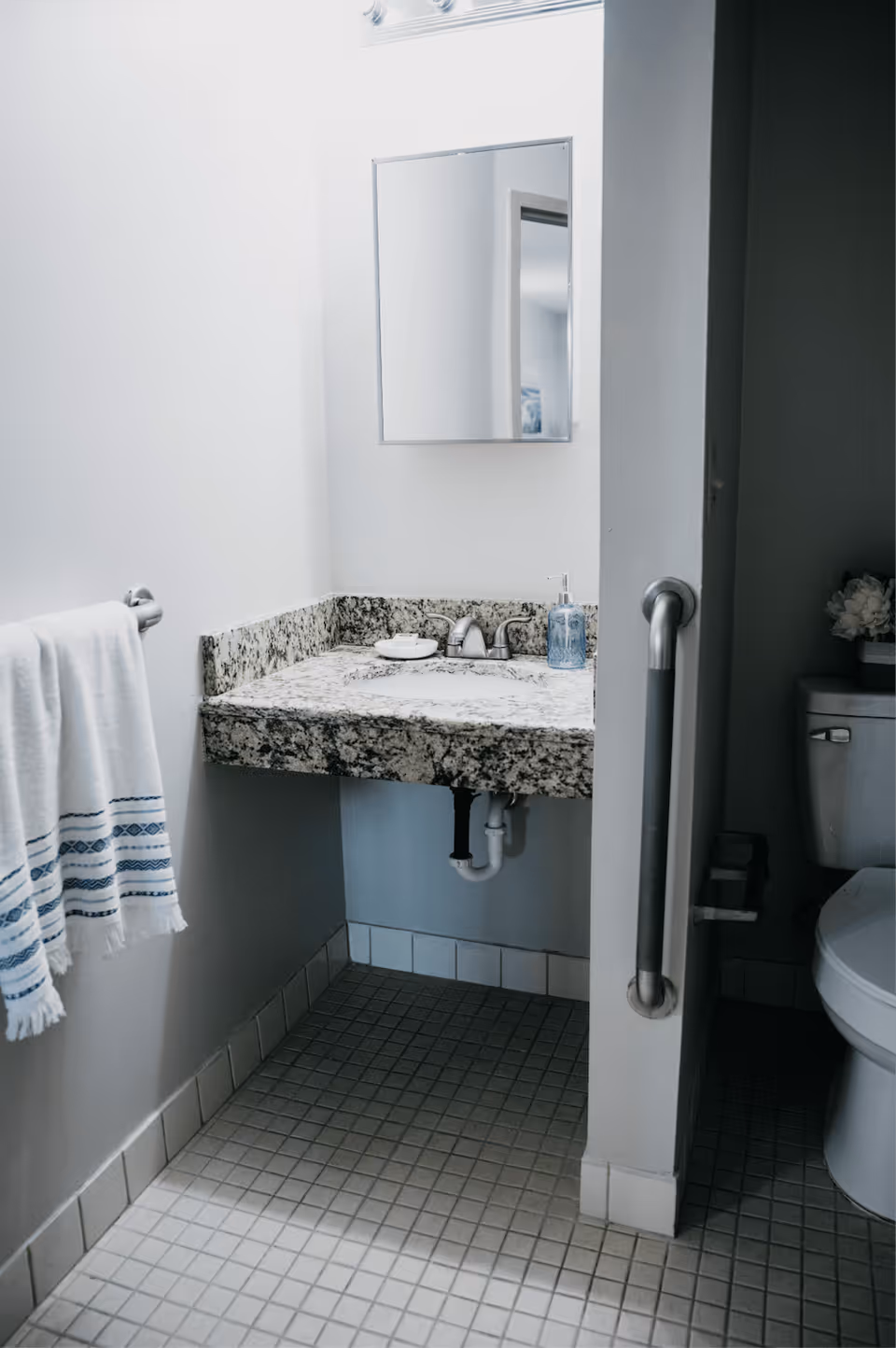 A bathroom with a granite countertop sink, a wall-mounted mirror above the sink, a towel rack with white towels featuring blue patterns, a toilet partially visible with a grab bar next to it, and tiled floor and walls.