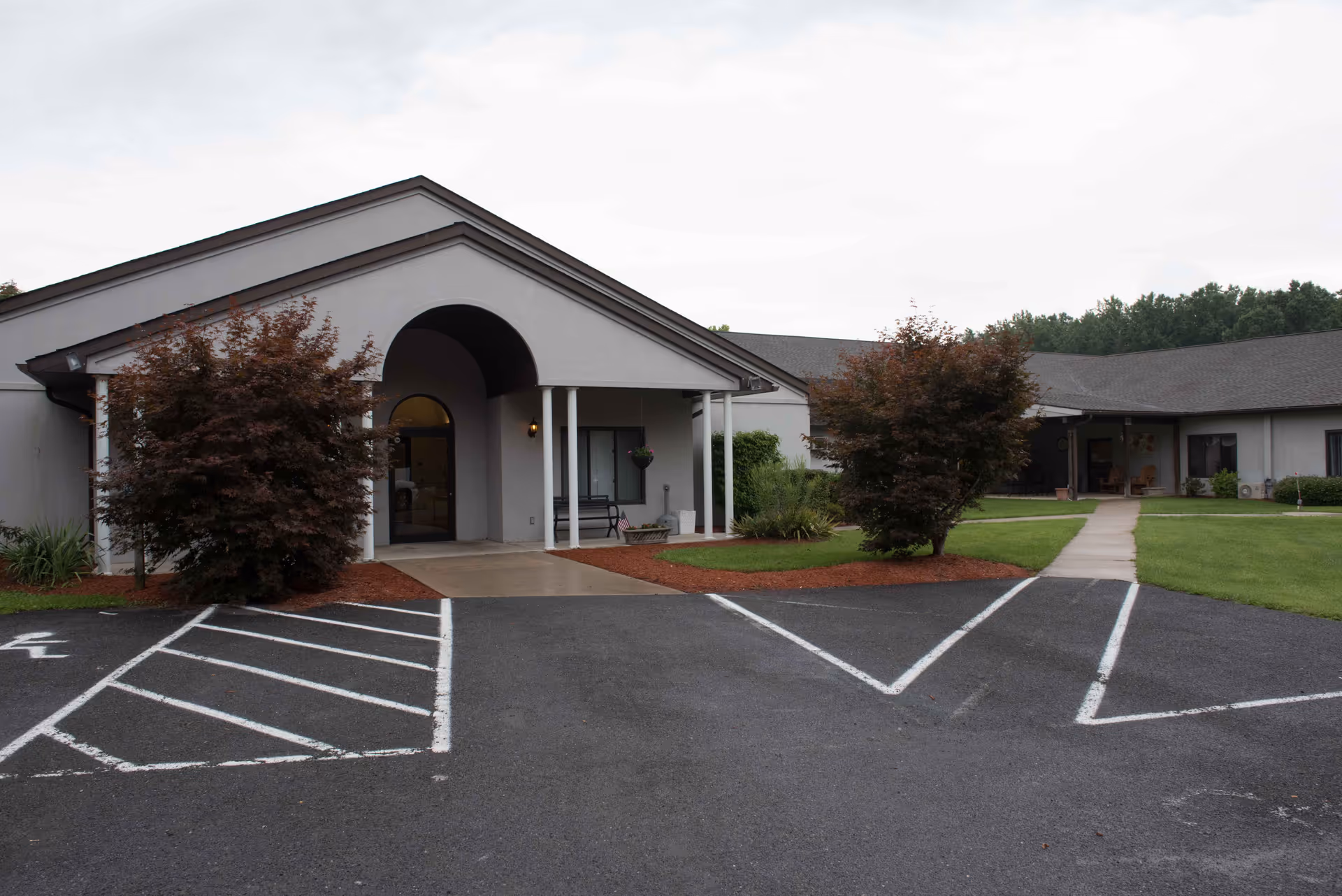 Exterior view of Berkeley Springs Healthcare Center showing a single-story building with a covered entrance supported by white columns. There are two small trees and landscaped areas with mulch and grass near the entrance. The parking lot in front has marked spaces including a handicapped spot.