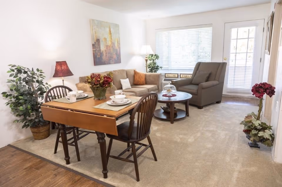 A cozy living room and dining area in a senior living facility featuring a small wooden dining table set for two with floral tableware, a beige sofa with cushions, a gray armchair, a round wooden coffee table with a glass dome, potted plants, a floor lamp, and a large window with blinds letting in natural light.
