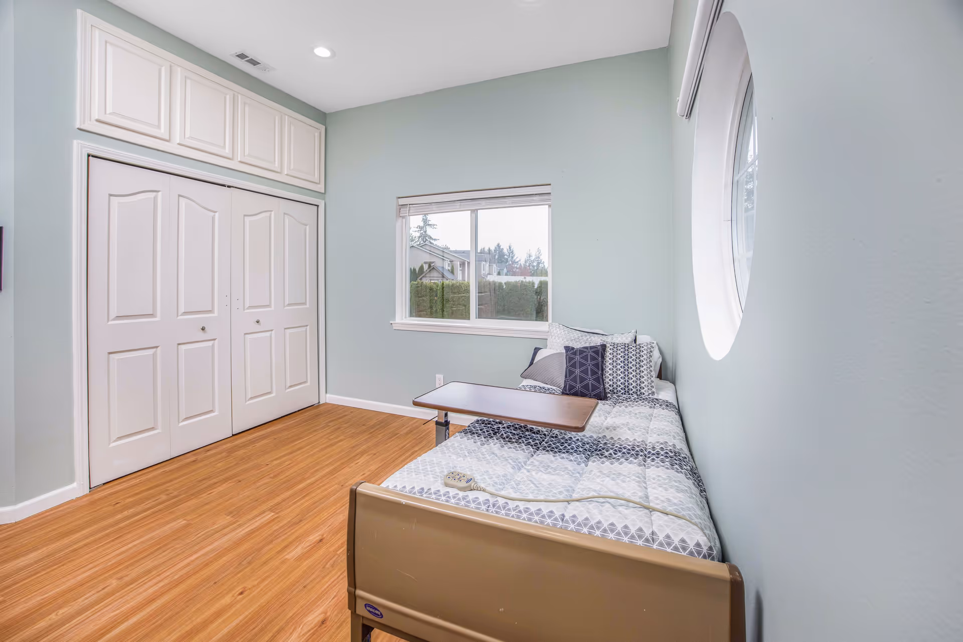 Small bedroom with a single bed against a pale green wall, a round window, a rectangular window, and double closet doors.
