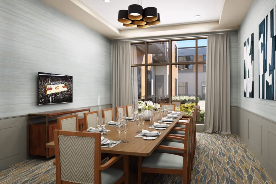 A dining room with a long wooden table set for a meal with plates, napkins, glasses, and silverware. There are twelve wooden chairs with cushioned seats around the table. A large window with curtains lets in natural light and shows an outdoor courtyard with plants. A flat-screen TV is mounted on the wall showing a basketball game, and modern artwork is displayed on the opposite wall. The room has a patterned carpet and a contemporary ceiling light fixture.