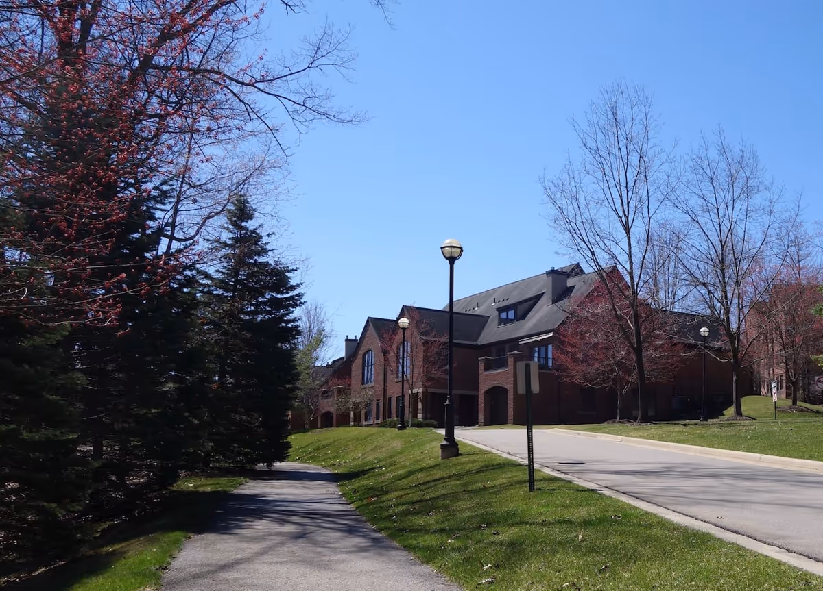 Brick multi-story building set behind a driveway and a tree-lined walkway with lampposts under a clear blue sky.