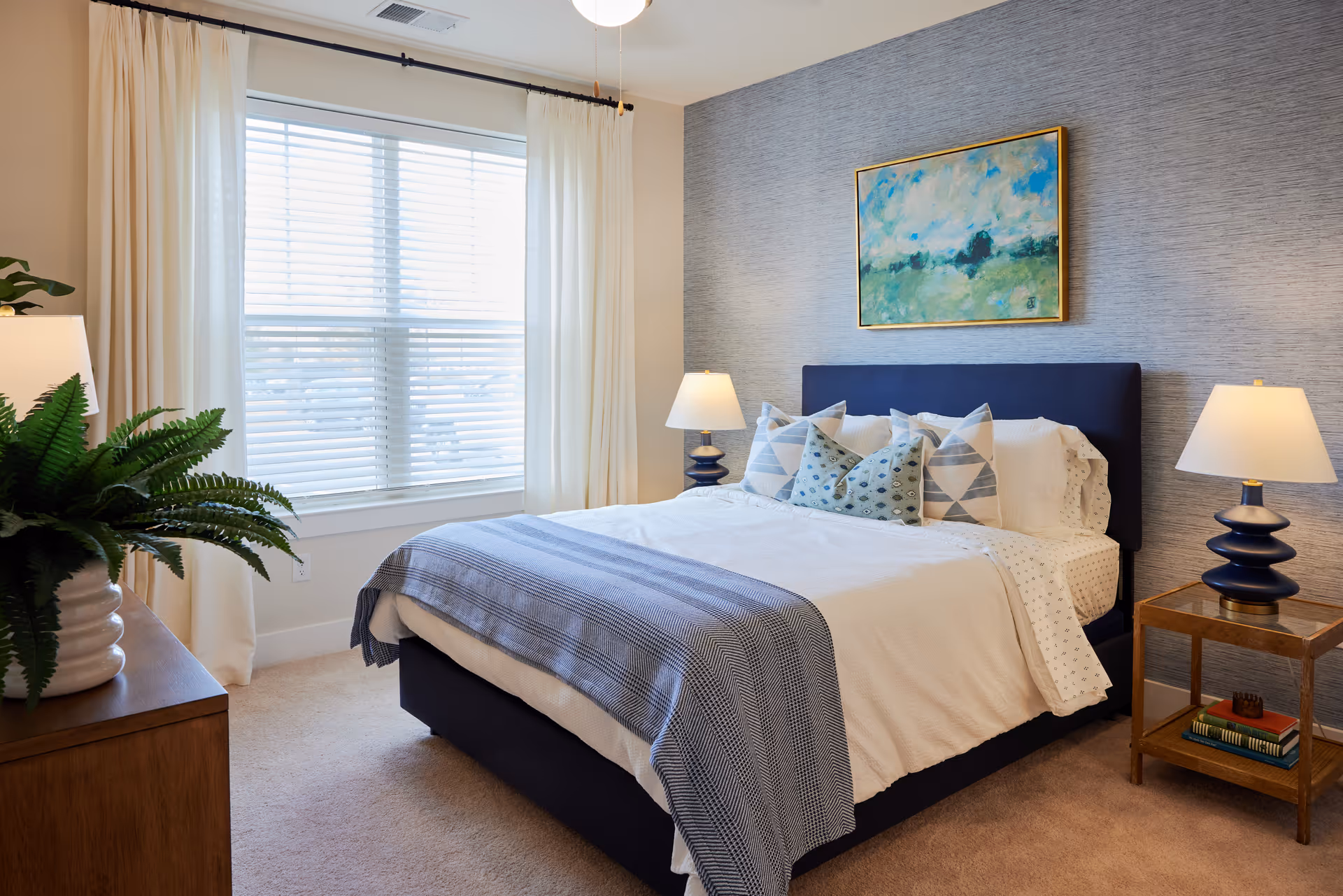 Bright, furnished bedroom featuring a bed with patterned pillows, two bedside lamps, a window with curtains, and wall art.