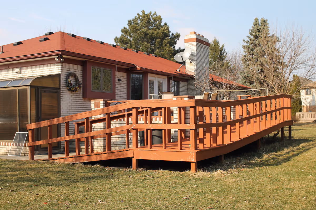 Exterior view of a single-story brick building with a red roof and a wooden wheelchair-accessible ramp leading to a patio area with outdoor chairs. Trees and grass surround the building under a clear sky.