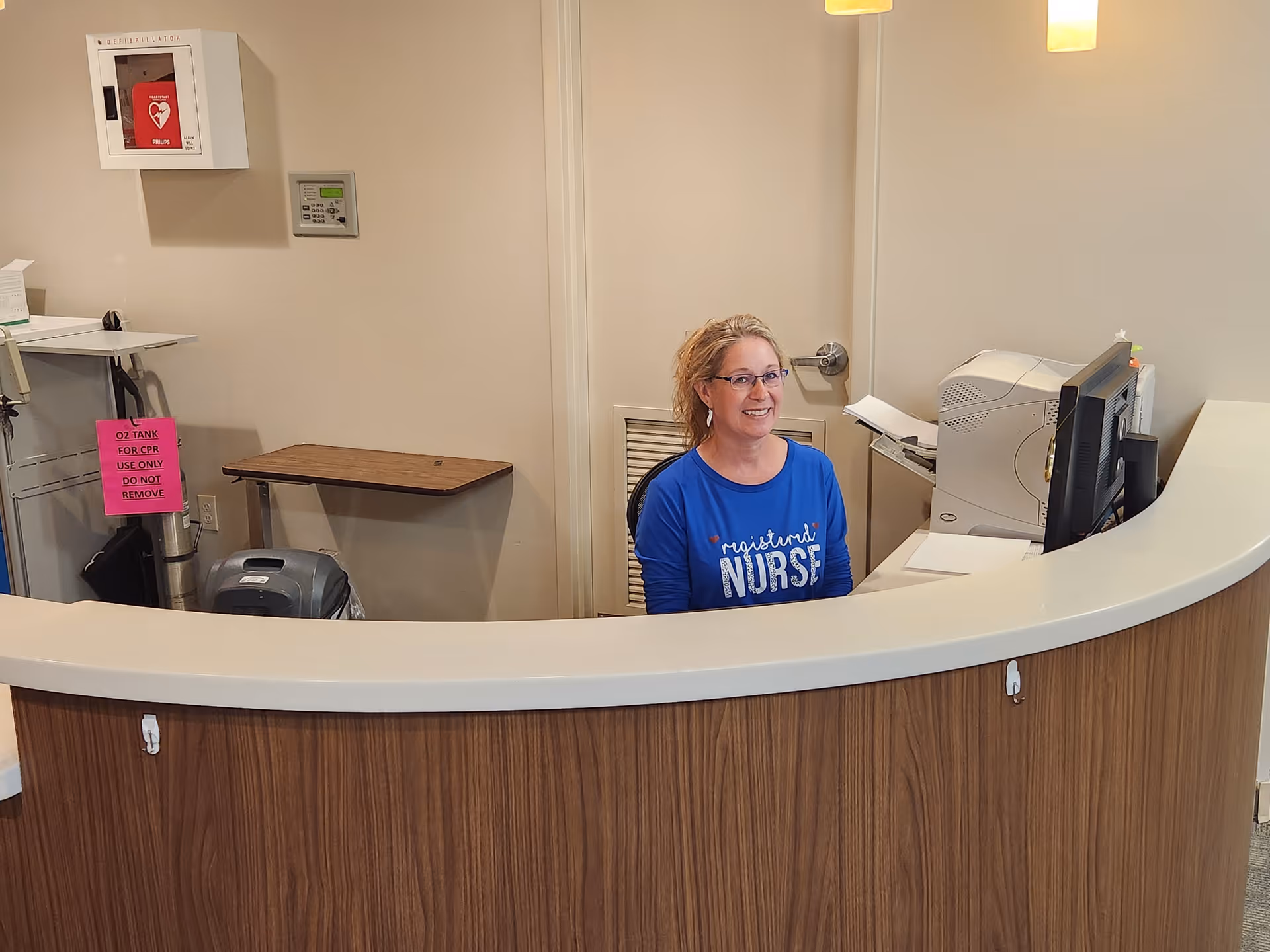 A woman wearing glasses and a blue shirt with the words 'registered NURSE' sits behind a curved reception desk in an office setting. Behind her is a computer, printer, and a closed door. On the wall to the left, there is a mounted emergency defibrillator and a sign indicating an oxygen tank for CPR use only.
