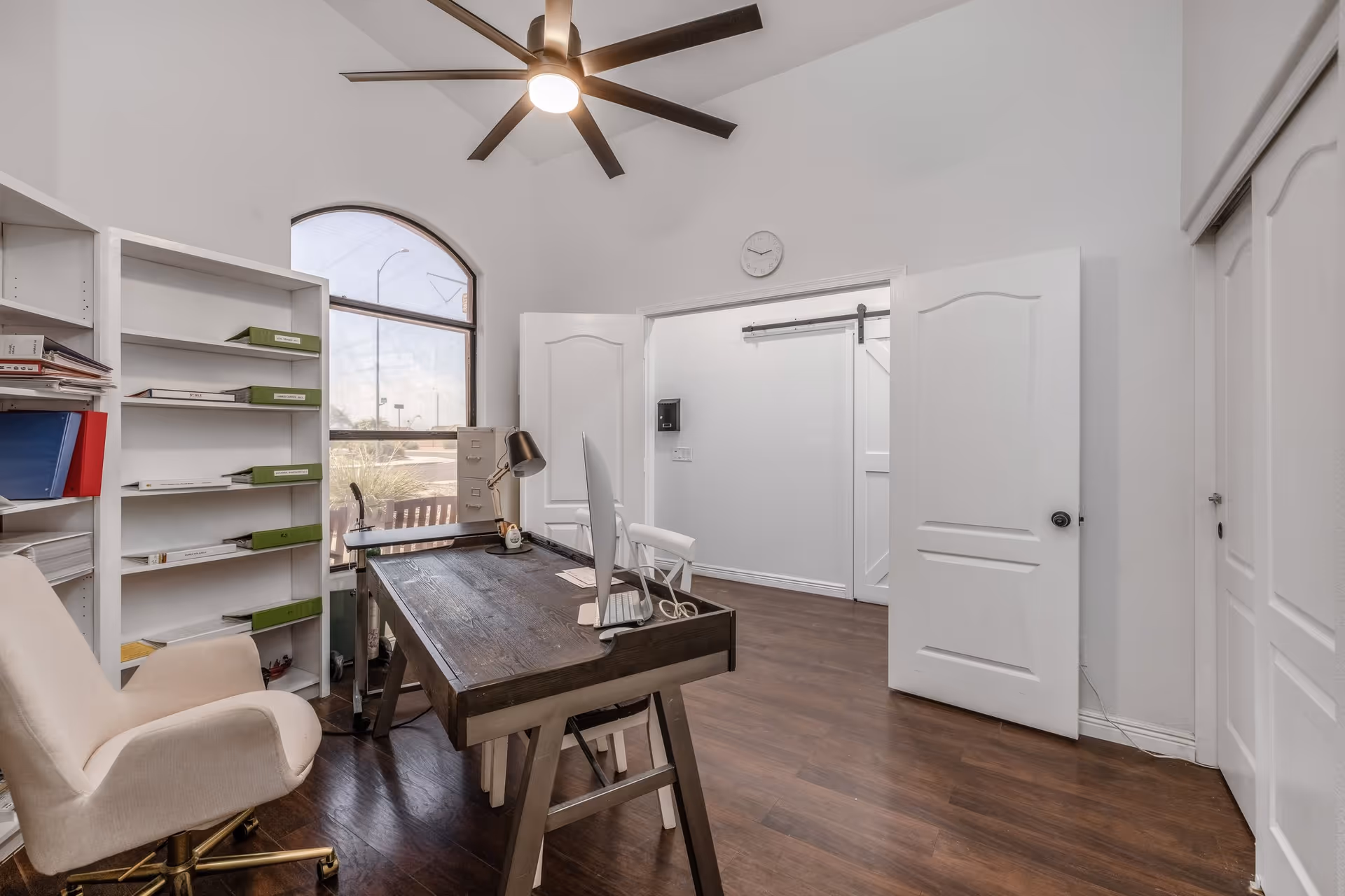 Bright interior office with a wooden desk, swivel chair, shelving, a large arched window and a ceiling fan.