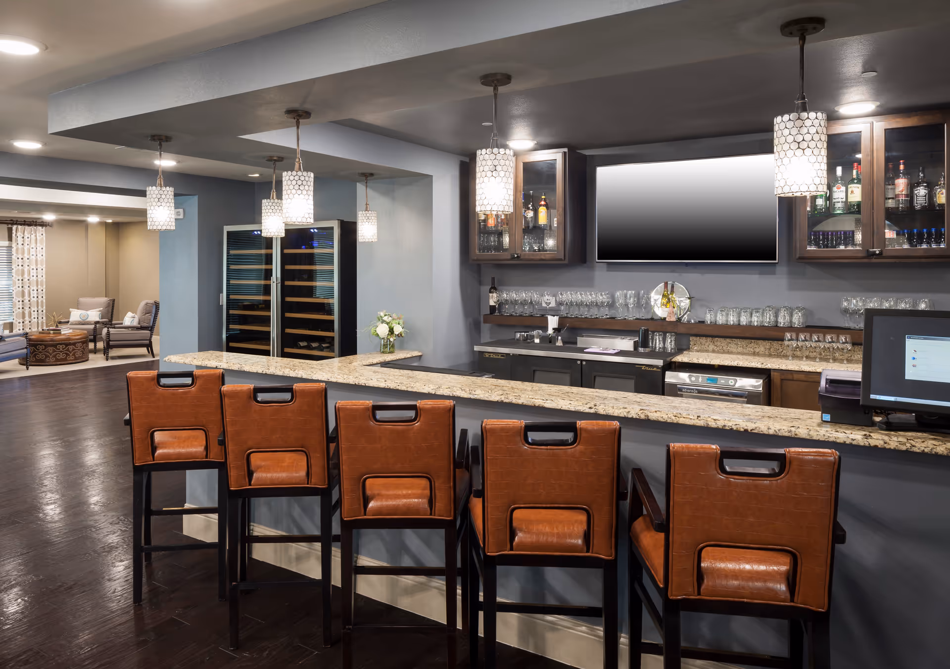 Interior view of a modern bar area with a granite countertop and six brown leather bar stools. Behind the bar are glass cabinets with bottles and glasses, a large flat-screen TV mounted on the wall, and a wine cooler. Pendant lights hang from the ceiling, and a seating area with chairs and a round ottoman is visible in the background.
