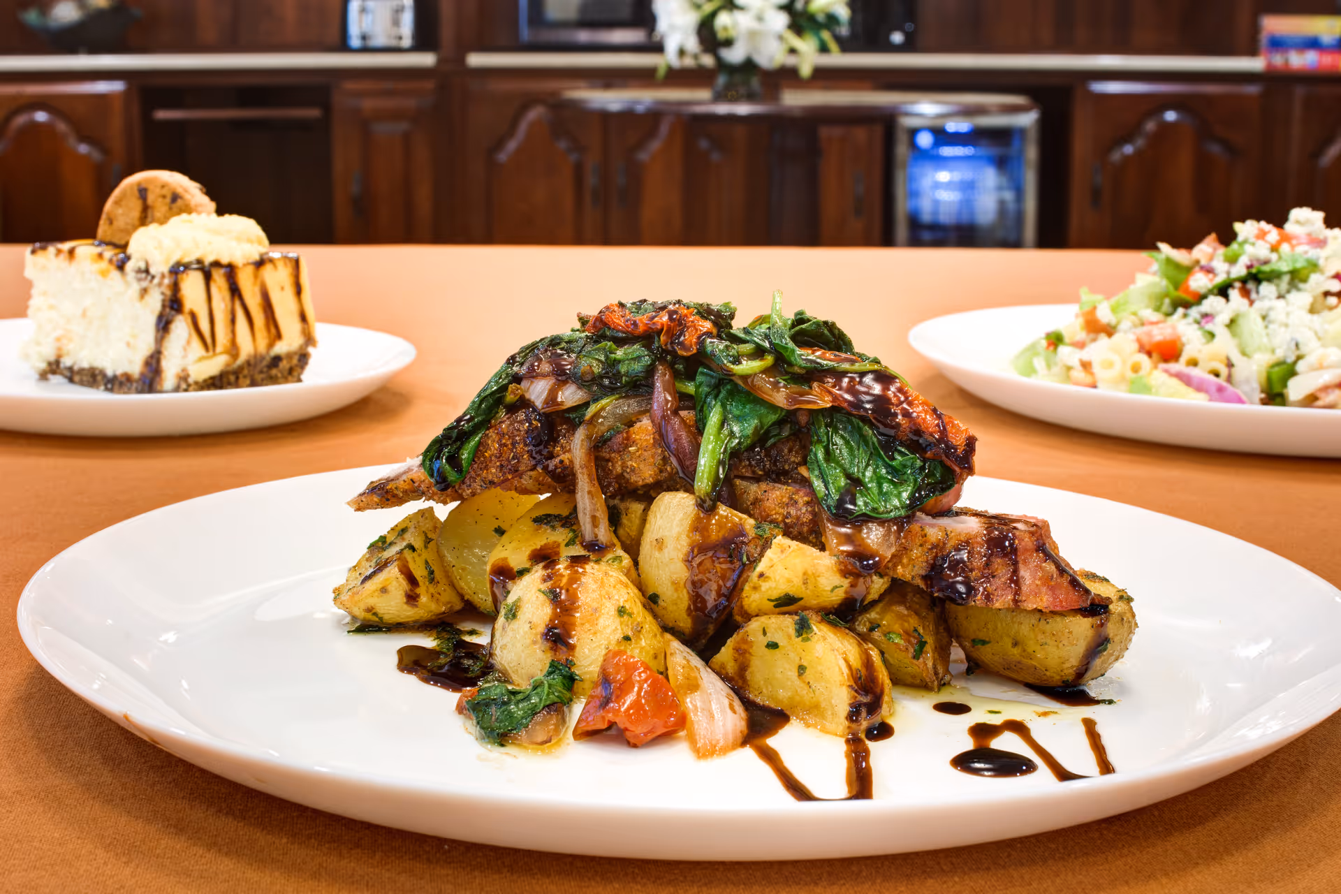 A plated meal featuring roasted potatoes topped with sautéed greens and grilled meat, drizzled with a dark sauce, with a slice of cheesecake and a salad in the background on a brown tablecloth.