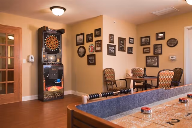 A communal game room showing a shuffleboard table in the foreground, a dart machine against the wall, and a small table with chairs beneath framed photos.