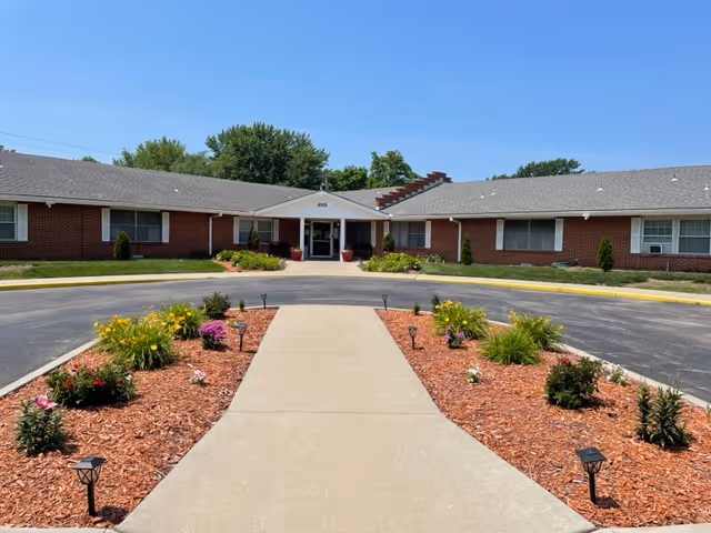 Front exterior view of Heritage Avonlea of Olathe, showing a single-story brick building with a central entrance, surrounded by landscaped flower beds and a circular driveway.