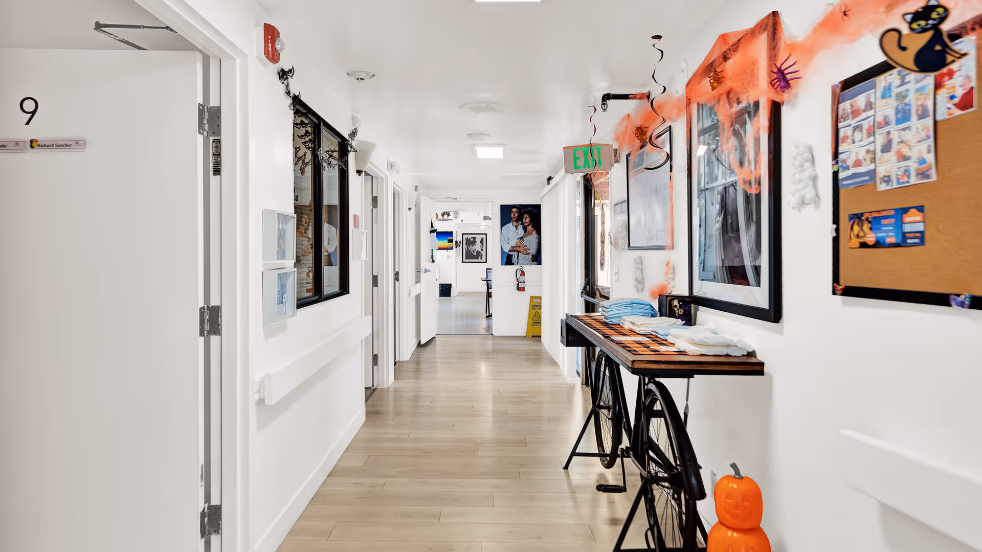 A bright hallway in a senior living facility decorated with Halloween-themed items including orange spider webs, hanging black bats, and a small pumpkin decoration on the floor. The hallway has light wood flooring, white walls, and several doors on the left side. A bicycle-themed table with folded towels is positioned along the right wall, which also features framed pictures and a bulletin board with various notices and photos. An exit sign is visible at the end of the hallway.