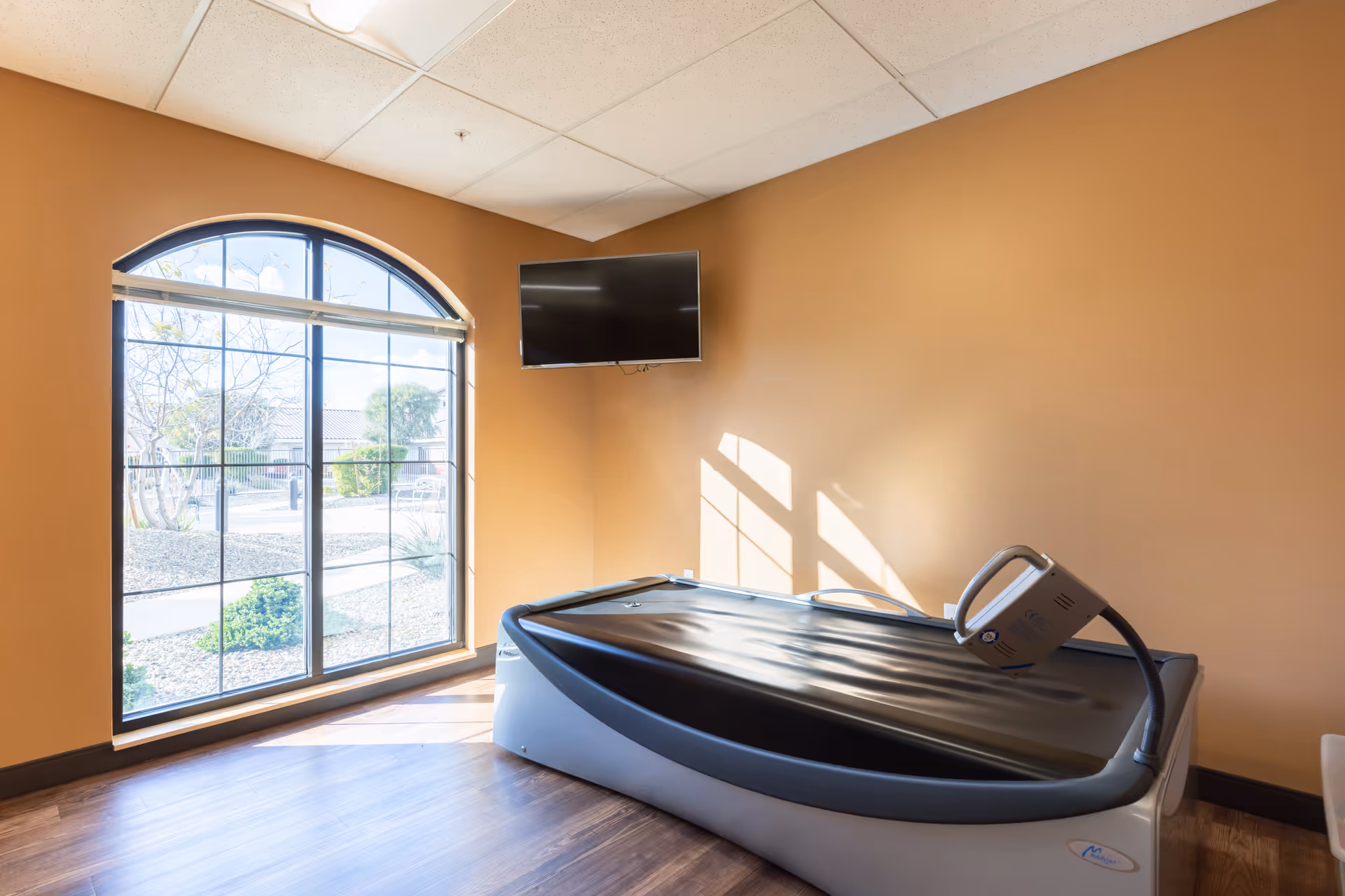 A therapy or treatment room with a large arched window letting in natural light, a mounted flat-screen TV on the wall, and a modern therapy or massage bed with a control device on it. The walls are painted a warm beige color and the floor is wood.