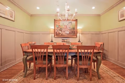 A dining room with a wooden dining table surrounded by six wooden chairs with cushioned seats. The room has light green walls with beige wainscoting and a decorative chandelier hanging above the table. There is a large framed painting on the wall behind the table, flanked by two table lamps on a sideboard. The floor is wooden with a patterned area rug under the table.