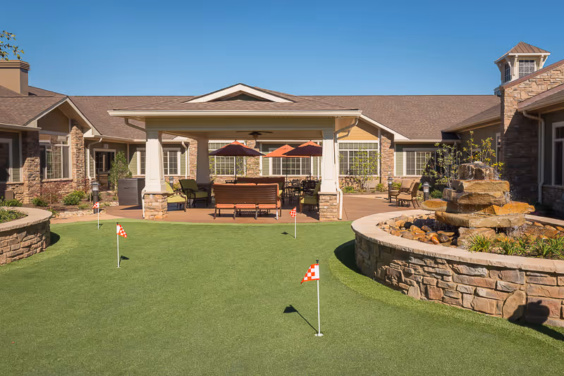 Outdoor courtyard area at a senior living facility with a putting green featuring small red and white checkered flags, a covered seating area with chairs and tables under umbrellas, and a stone water fountain surrounded by a stone wall. The building exterior is visible in the background under a clear blue sky.