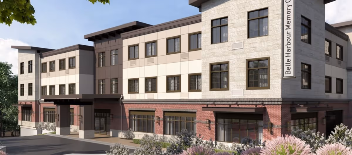 Exterior view of a modern three-story building with a mix of brick and light-colored paneling, large windows, and a covered entrance. The building has a sign that reads 'Belle Harbour Memory Care'. There are shrubs and plants in the foreground and a clear sky above.