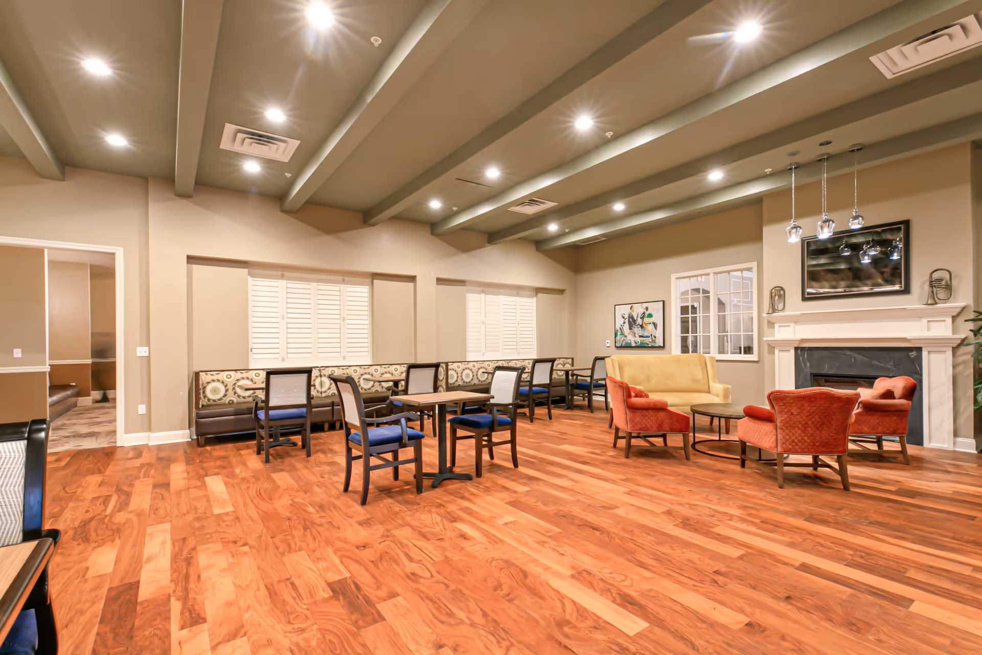 A spacious common area with wooden flooring, multiple tables with chairs, a yellow sofa, and three red armchairs arranged around a round coffee table in front of a white fireplace. The room has beige walls, a ceiling with exposed beams and recessed lighting, and a window with white shutters. There is also a framed artwork on the wall and pendant lights hanging above the fireplace.