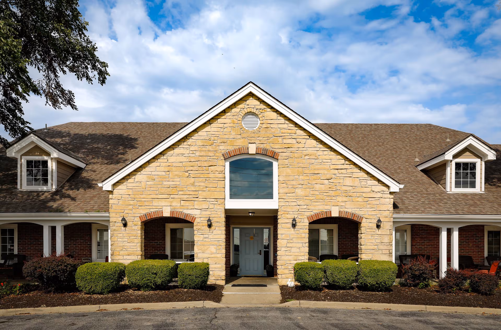Front exterior view of a single-story assisted living facility building with a stone facade, a central entrance door, symmetrical windows, and a gabled roof under a partly cloudy sky.