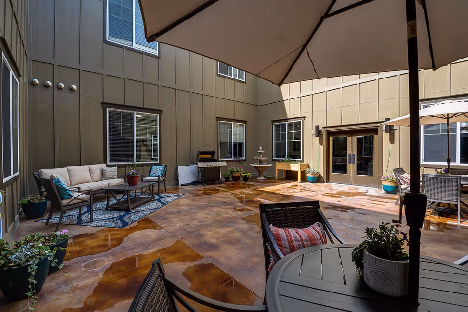 Outdoor courtyard area at Spring Gardens Senior Living Meridian featuring patio furniture including cushioned chairs and tables with umbrellas, potted plants, a decorative fountain, and a barbecue grill against the building's exterior walls.